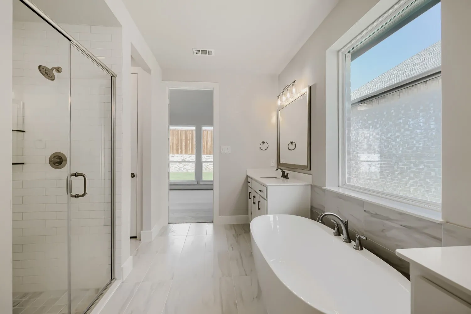 Contemporary bathroom featuring a freestanding soaking tub, glass-enclosed shower, dual vanity with quartz countertop, and large window with privacy film