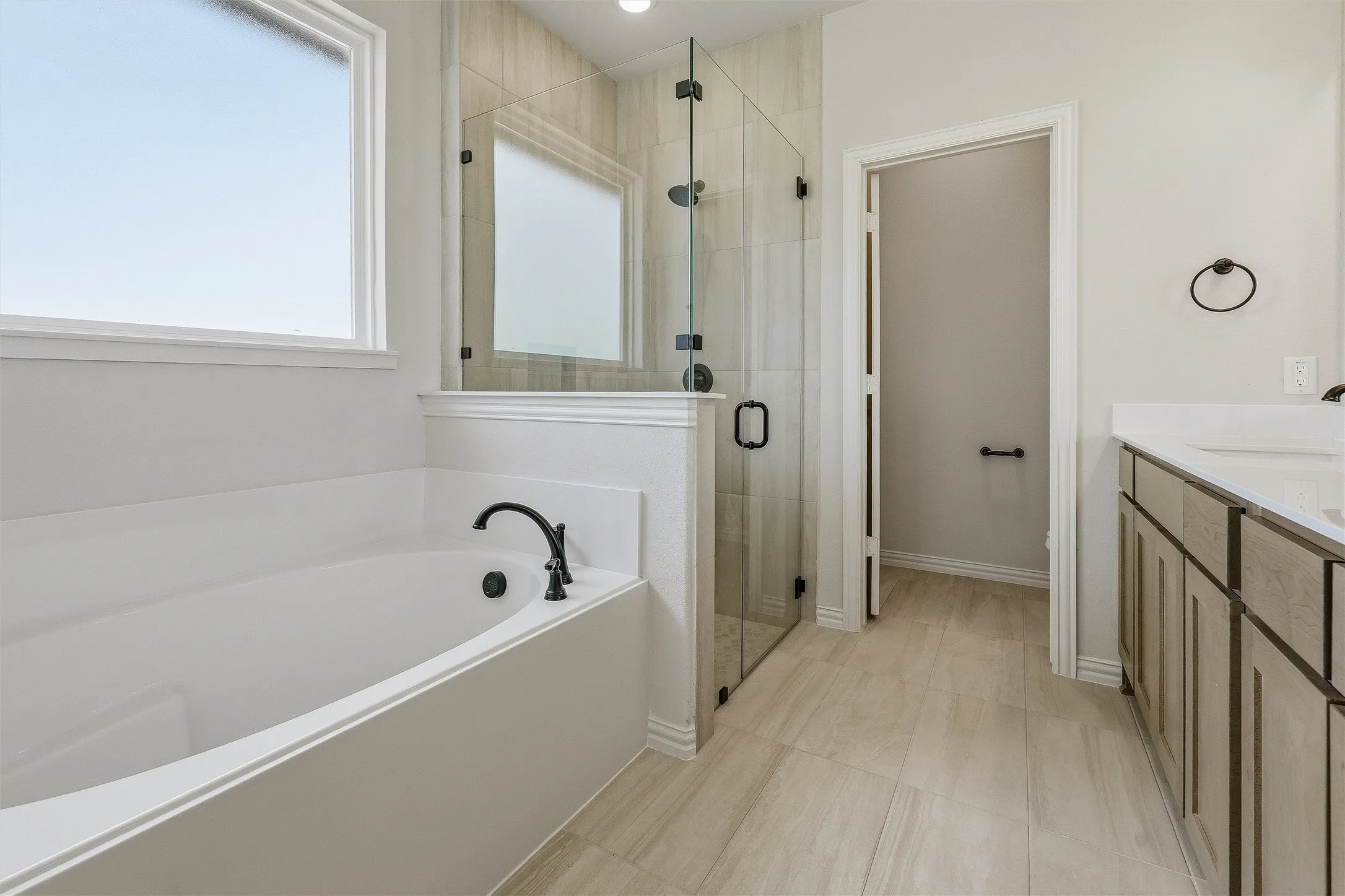 Spacious bathroom featuring a built-in soaking tub with matte black fixtures, a glass-enclosed shower, dual vanity with undermount sinks, and light-toned tile flooring