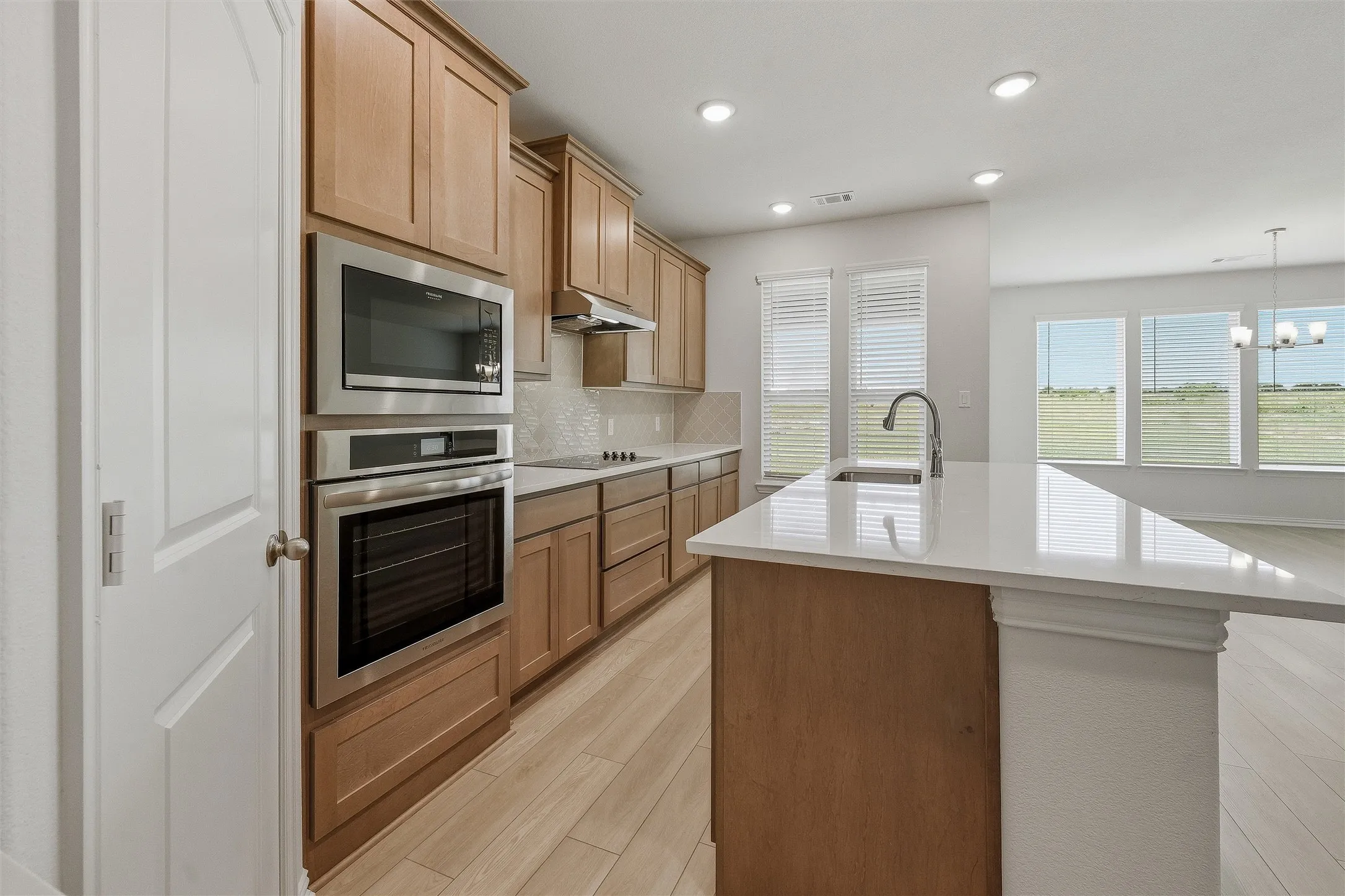 Kitchen featuring light wood-finish cabinetry, stainless steel appliances, a white tile backsplash, and a central island with a white countertop and integrated sink