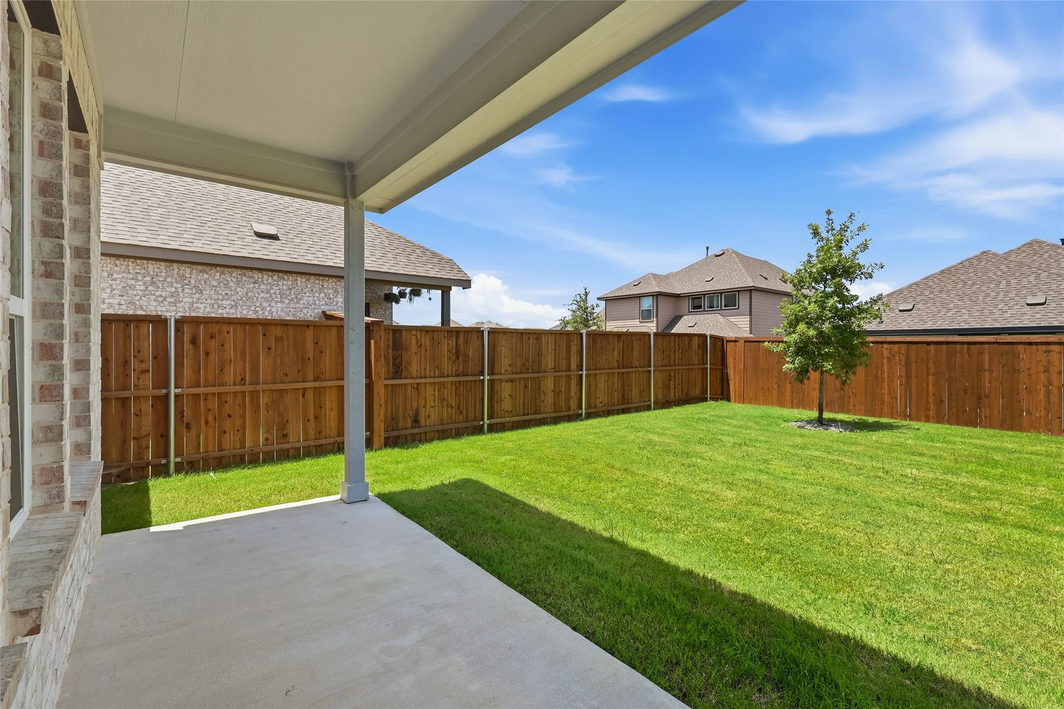 Expansive backyard featuring a covered patio, concrete slab, wood privacy fencing, and a manicured lawn with a young tree