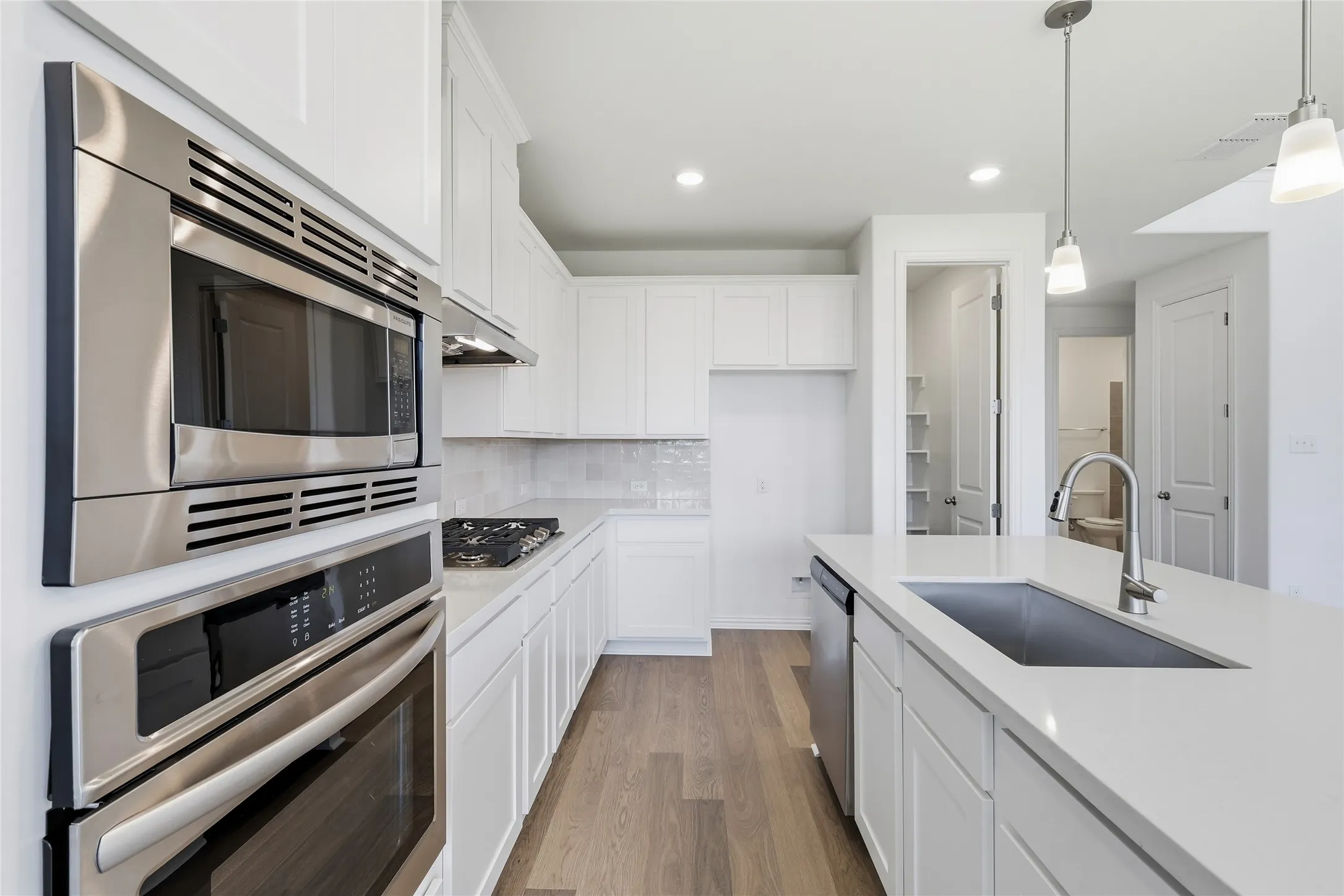 Modern kitchen featuring an island with an undermount sink and brushed nickel faucet, stainless steel appliances, white cabinetry, light countertops, and wood-finish flooring