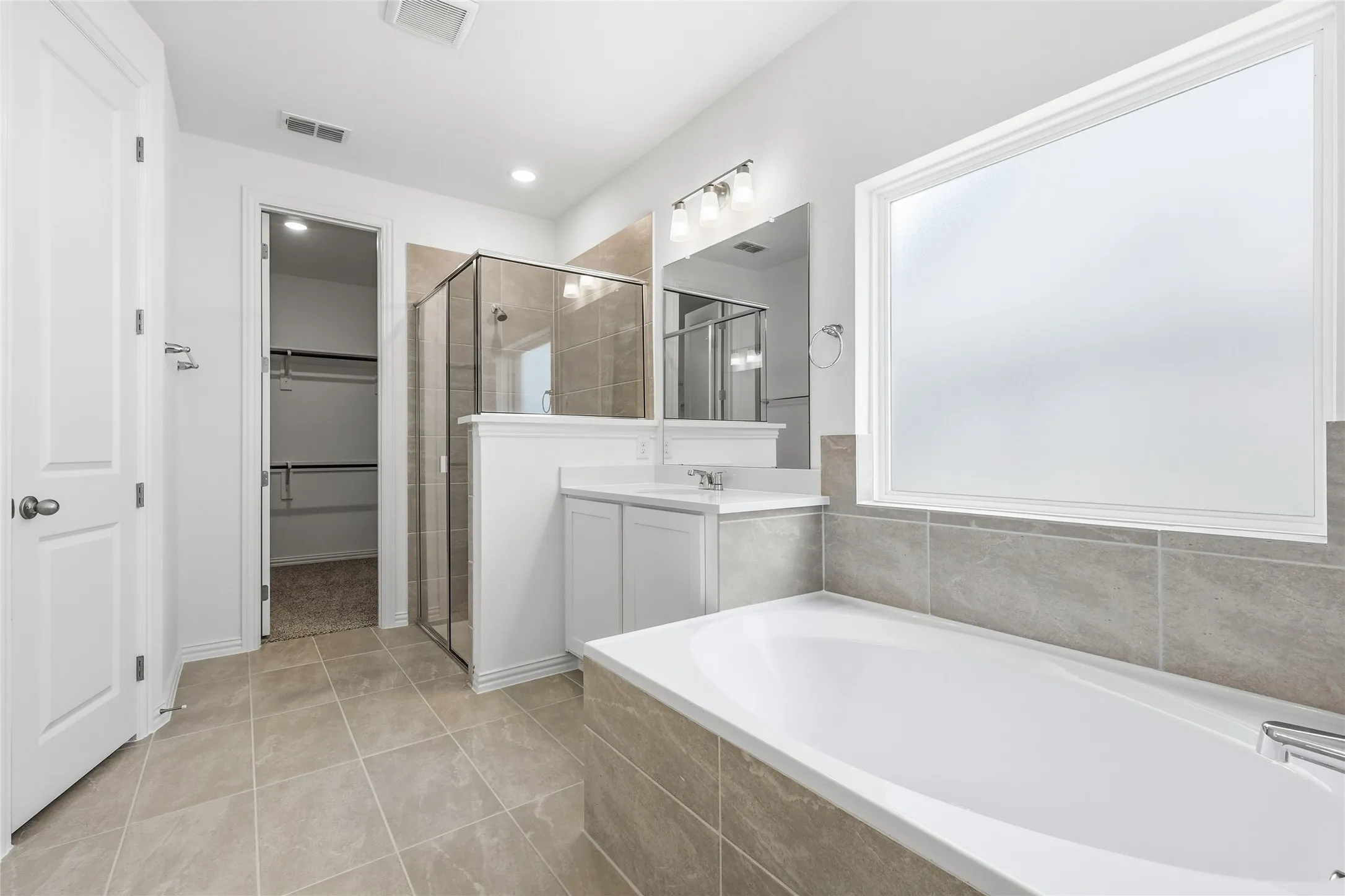 Spacious bathroom featuring a built-in soaking tub with tiled surround, a separate glass-enclosed shower, dual vanity with white cabinetry and quartz-finish countertops, and a large frosted window