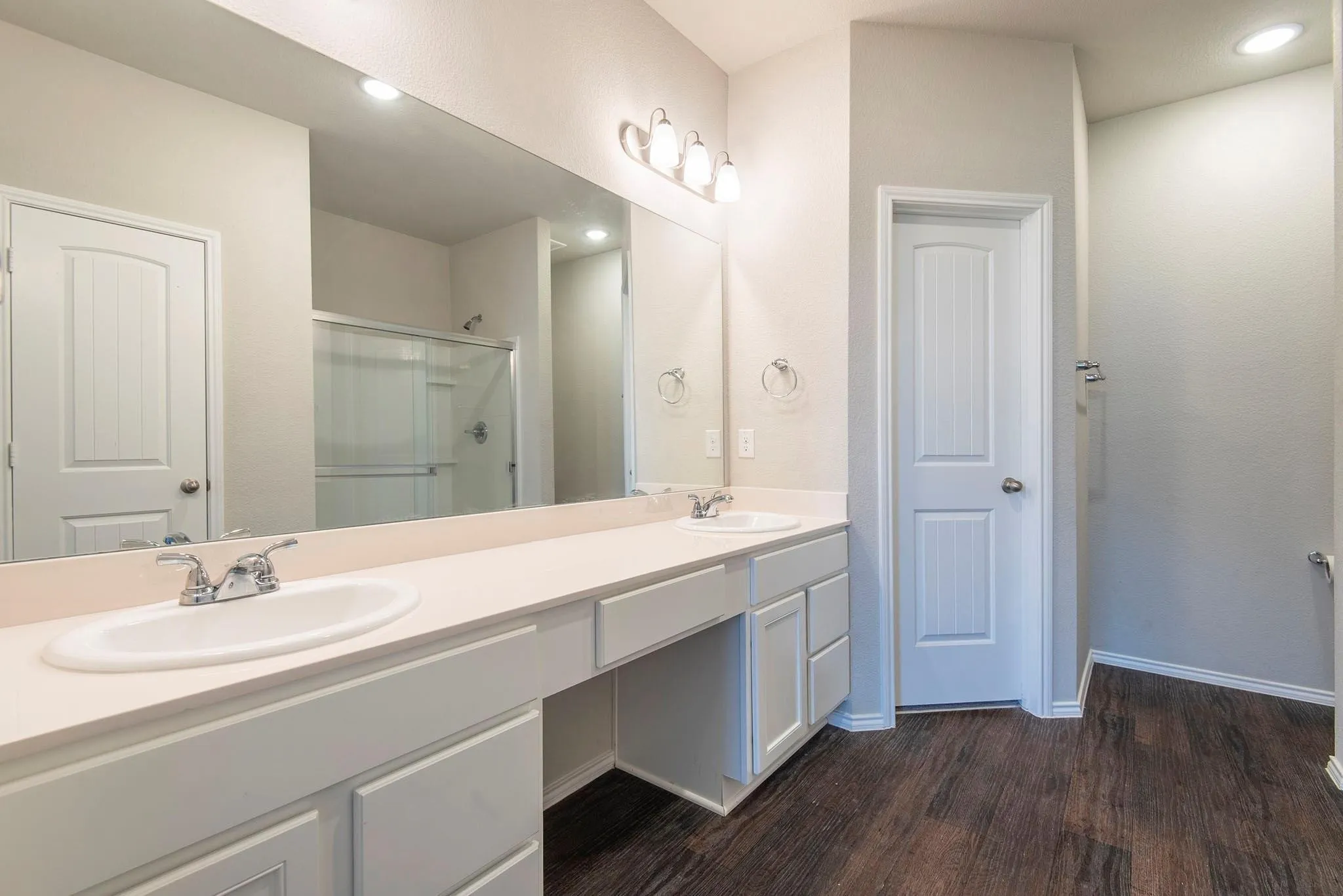 Dual vanity bathroom featuring an expansive countertop with dual sinks, abundant storage cabinetry, large framed mirror with integrated lighting, wood-finish flooring, and direct access to adjacent spaces through multiple doorways