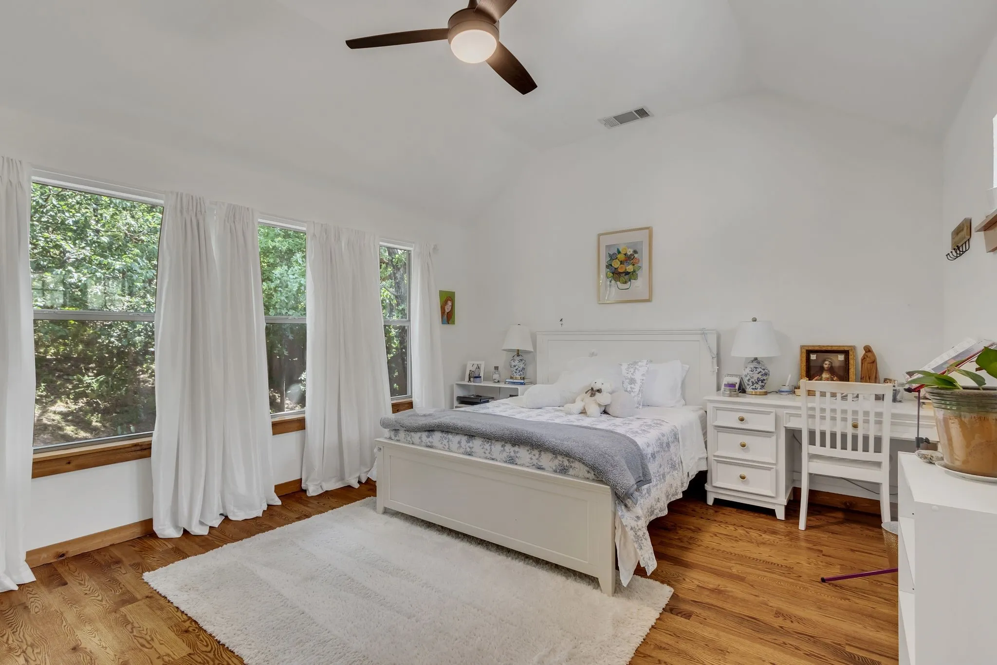 Vaulted ceiling bedroom featuring wood-finish flooring, a ceiling fan with integrated lighting, and three large windows with wood trim