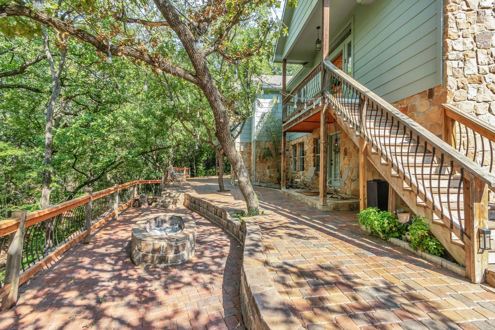Expansive brick paver patio featuring a built-in stone fire pit, wood railing with wrought iron balusters, a stone retaining wall, and mature trees