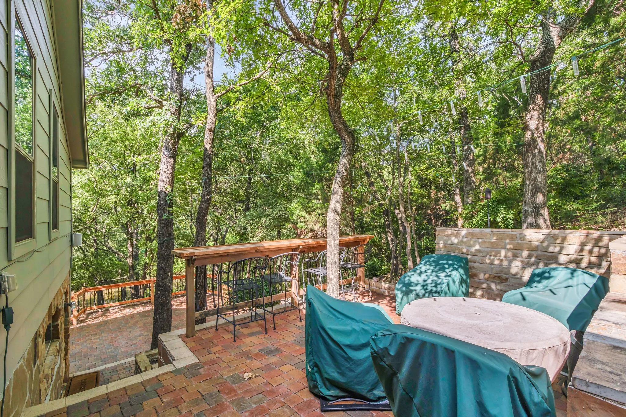 Brick paver patio surrounded by mature trees, featuring a built-in stone wall, a wood-finish bar, and a lower-level deck with wood railing