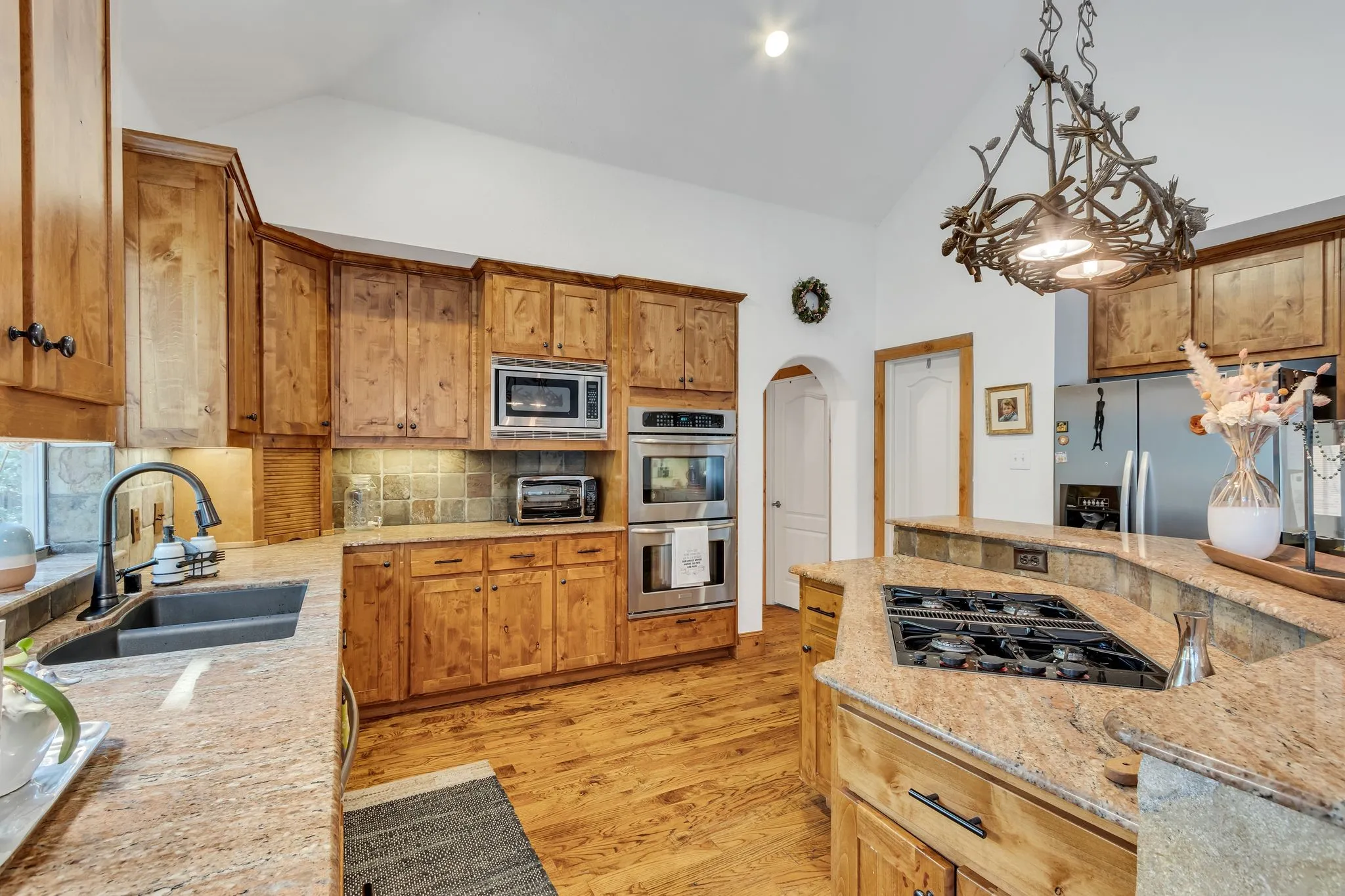 Kitchen featuring wood cabinetry, granite countertops, a built-in double oven, a gas cooktop on the island, and a vaulted ceiling with a decorative light fixture