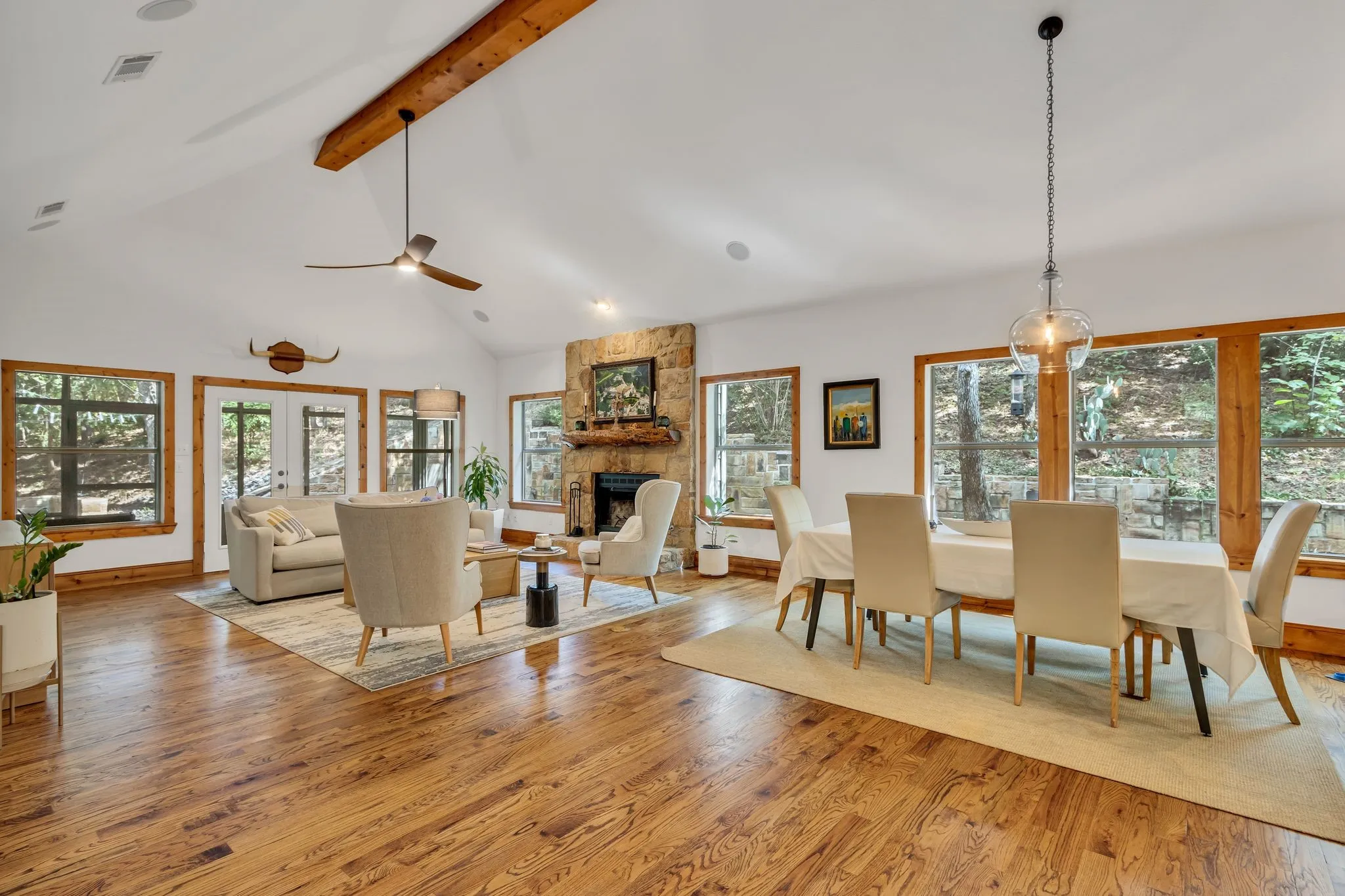 Spacious living area featuring high vaulted ceilings with exposed wood beams, a stone fireplace, wood-finish flooring, and multiple large windows with natural wood trim