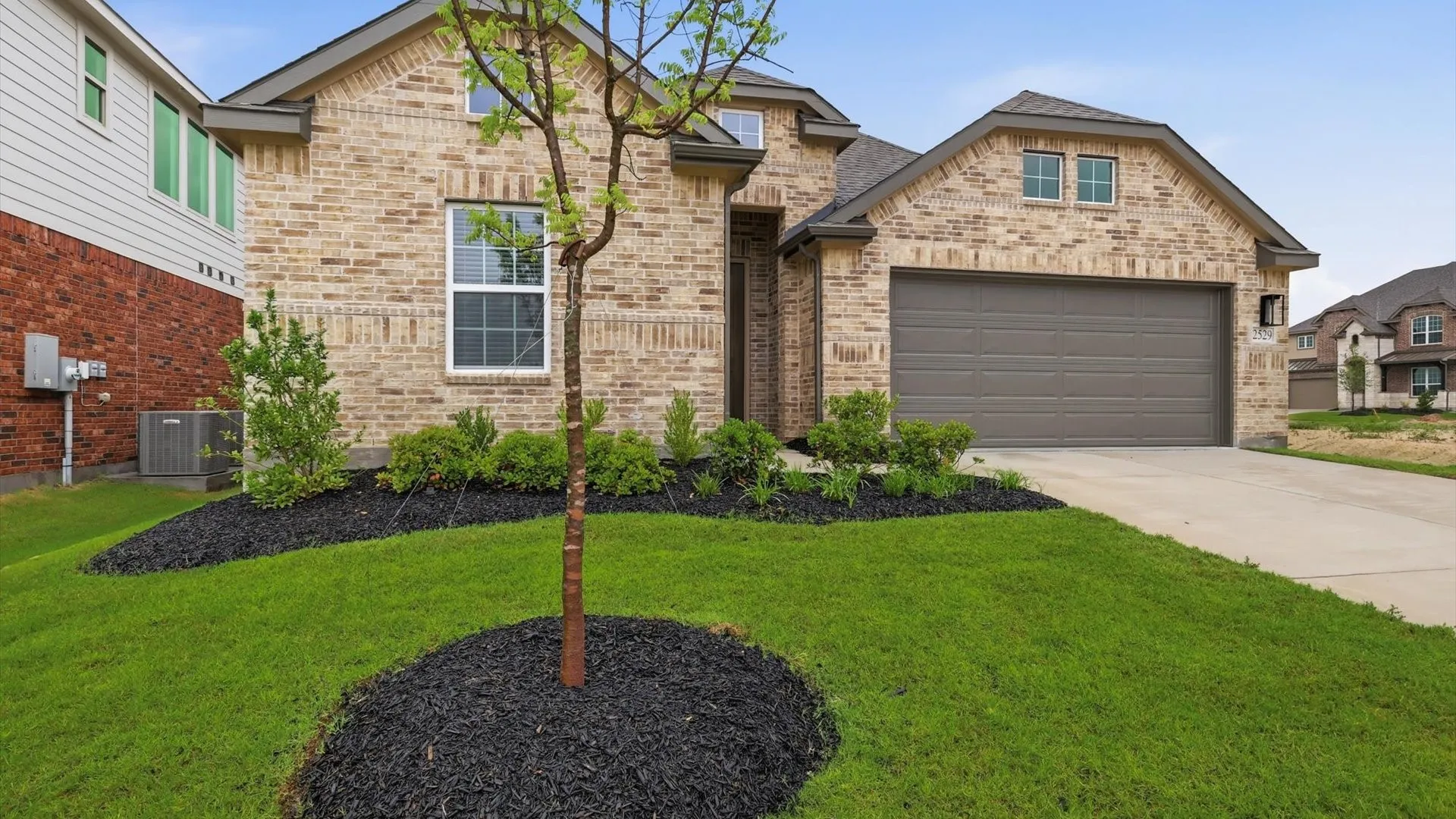 Brick exterior with an attached two-car garage, concrete driveway, and a manicured front lawn featuring a young tree and landscaped beds