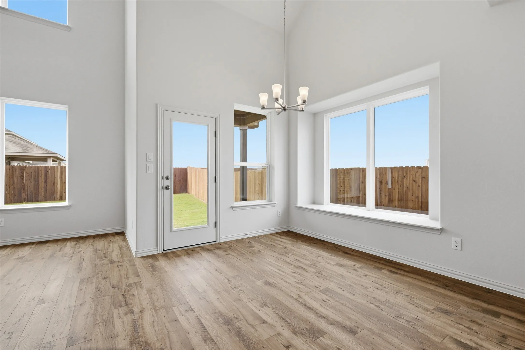 Spacious room with high ceilings, wood-finish flooring, multiple large windows, a glass-paneled door, and a contemporary chandelier