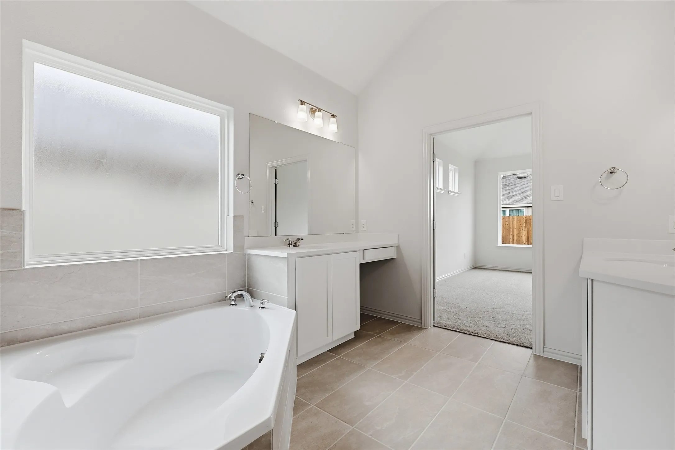 Contemporary bathroom featuring a built-in soaking tub with tile surround, a large frosted window, dual white vanities with integrated sinks, and neutral tile flooring