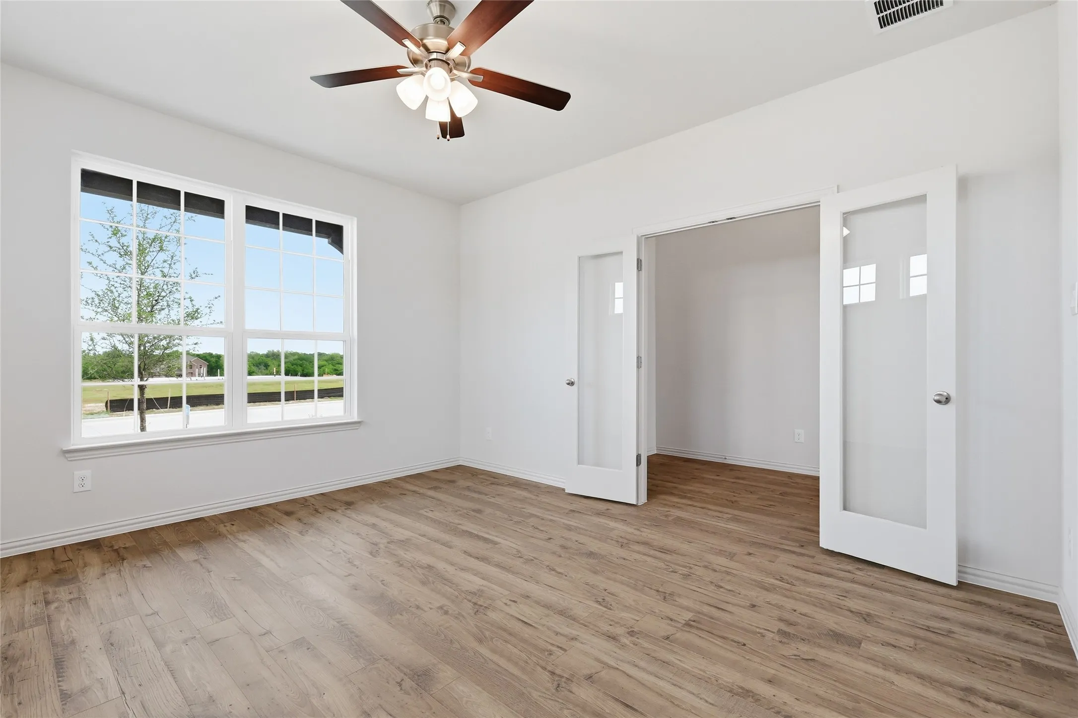 Spacious room with wood-finish flooring, a large multi-pane window, and a ceiling fan