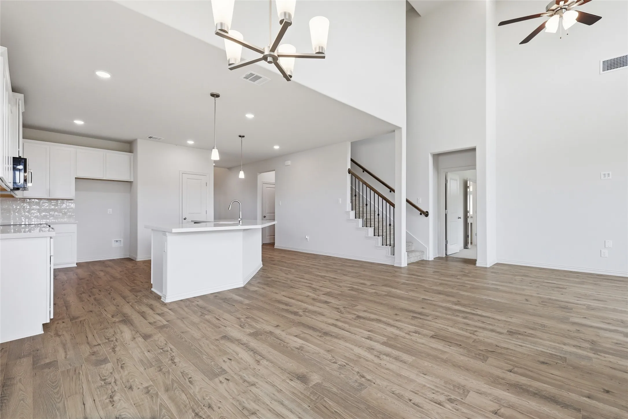 Open-concept living area featuring high ceilings, wood-finish flooring, a kitchen island with pendant lighting, white cabinetry, and a staircase with wrought iron balusters