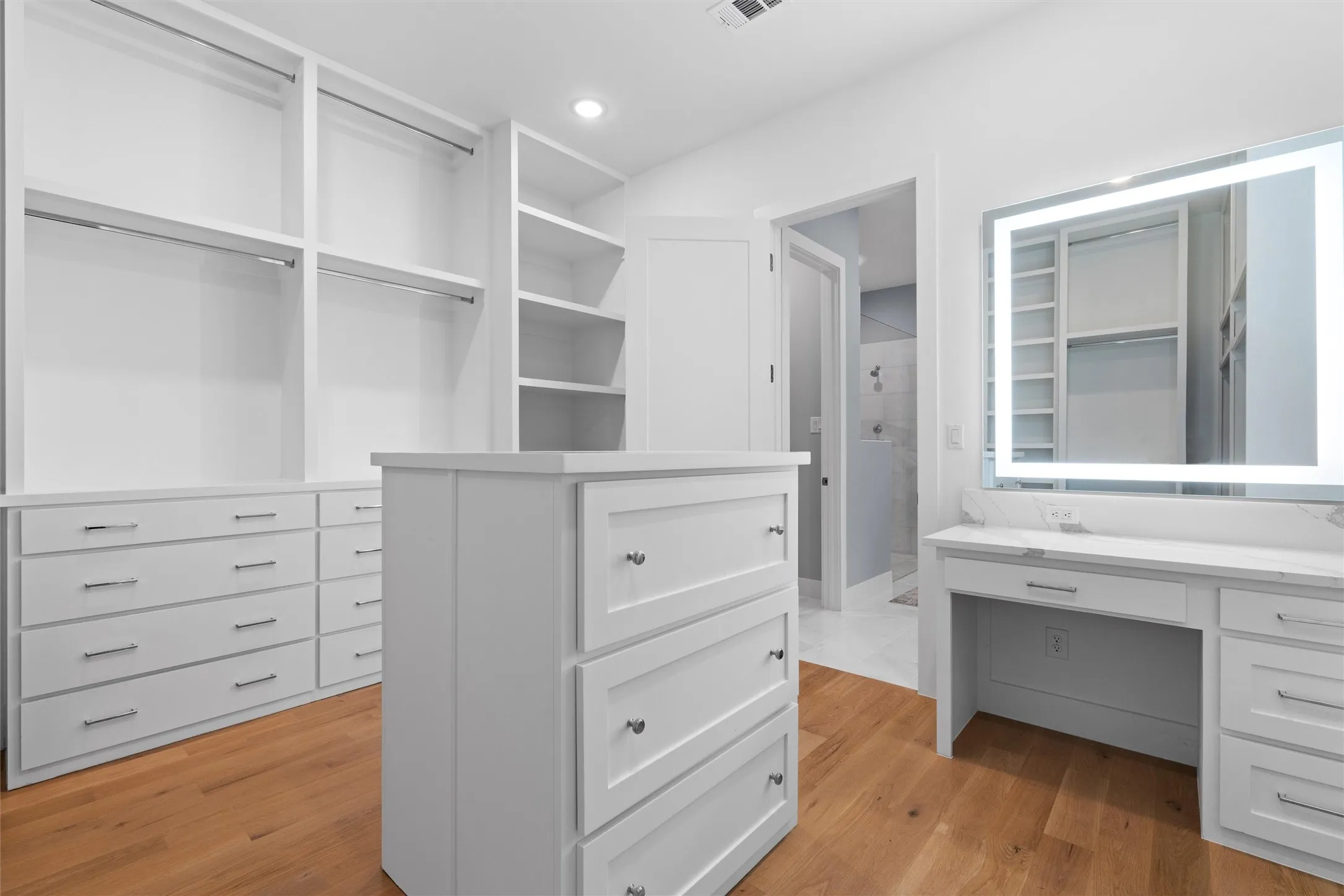 Master closet featuring light wood-type flooring, island of drawers, and lighted makeup mirror.