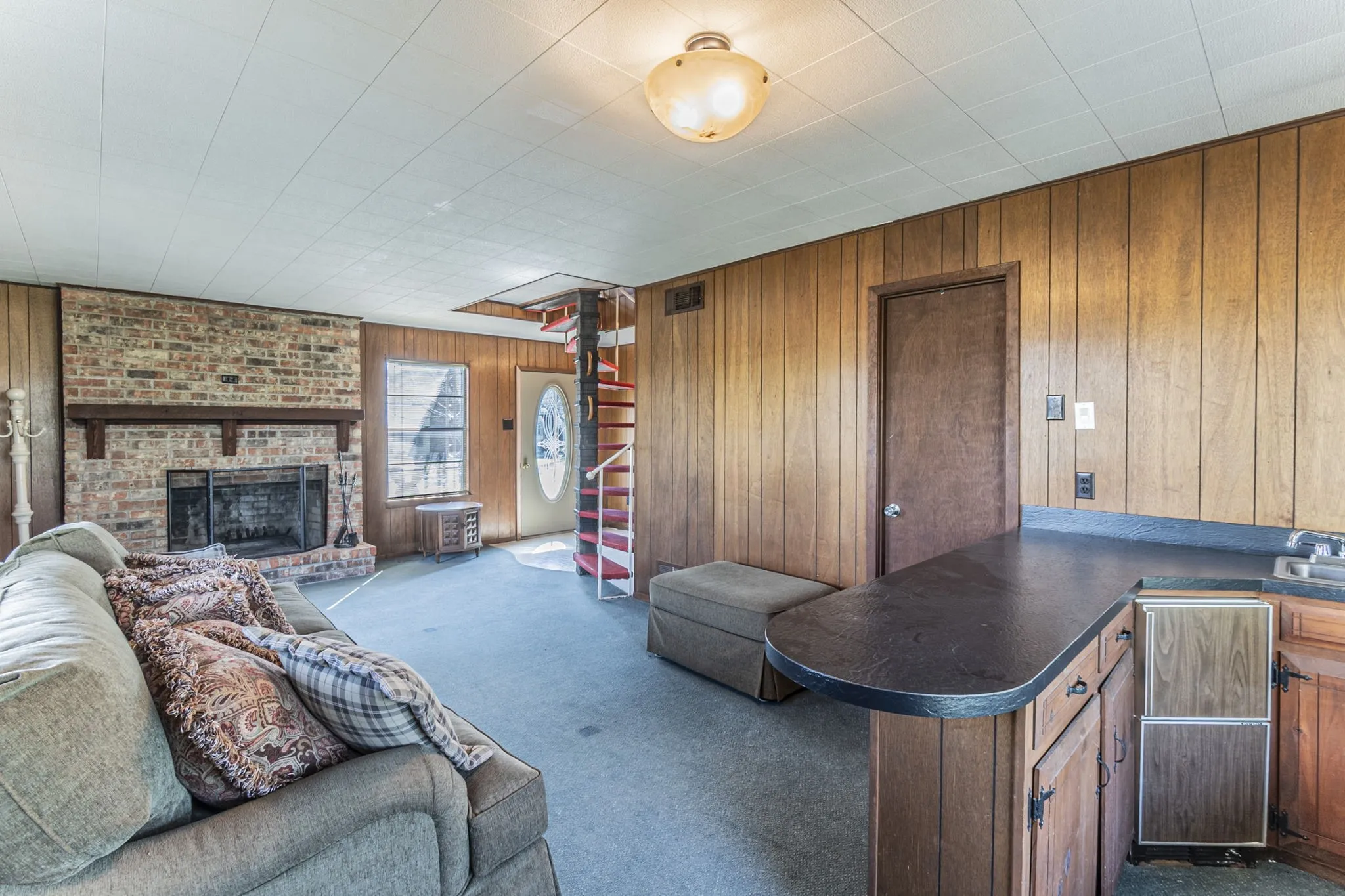 A view of the downstairs living room from the wet bar area. The front door is at the rear of this photo. Fireplace includes wood mantel and brick hearth.