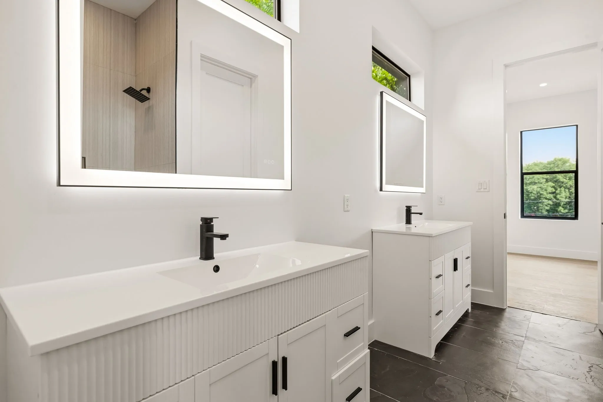 Dual vanity bathroom featuring backlit mirrors, matte black fixtures, crisp white cabinetry with fluted details, and dark tile flooring