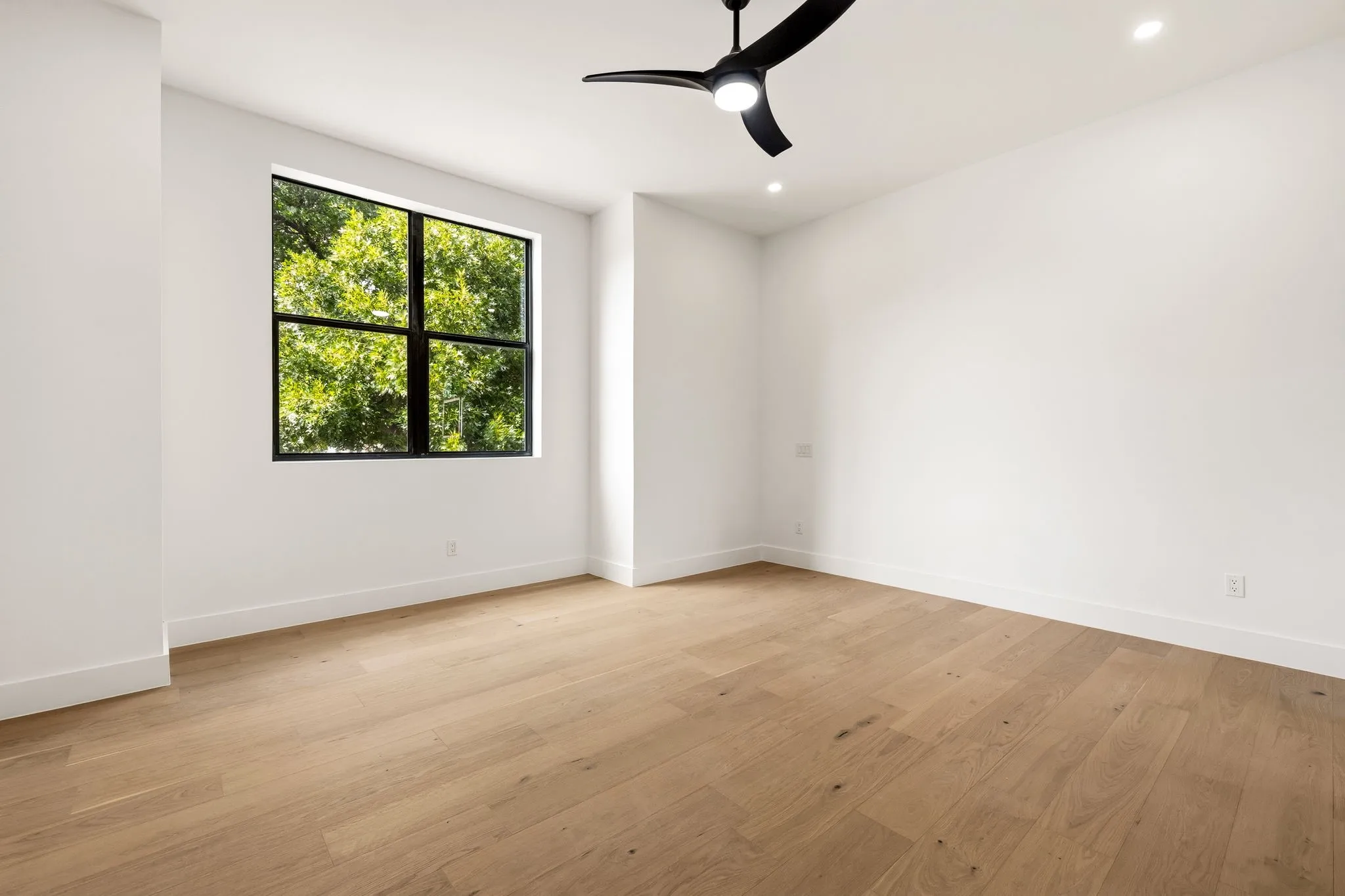 Spacious room featuring light wood-finish flooring, a large black-framed window, a modern black ceiling fan, recessed lighting, and crisp white walls