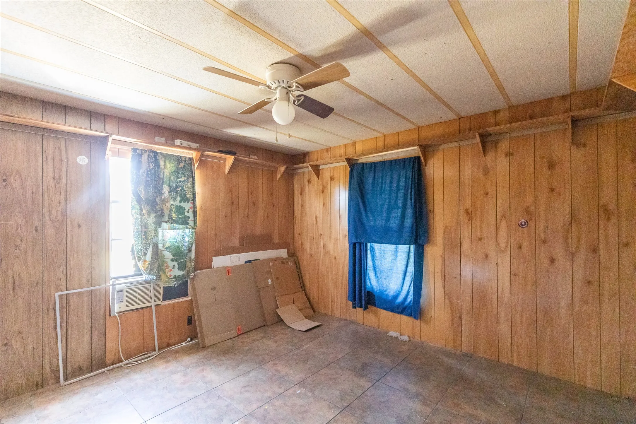 Room featuring wood paneling, tiled flooring, two windows, built-in shelving, and a ceiling fan with light fixture