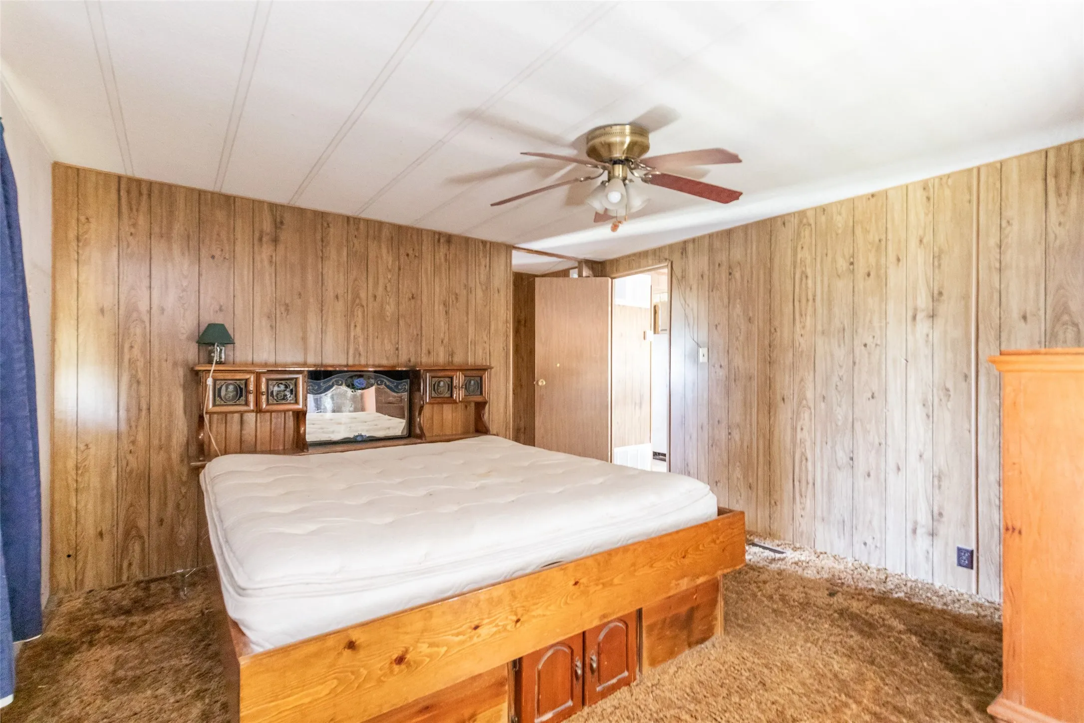 Wood-paneled wall, light wood-finish built-in headboard, wood-finish flooring, white paneled ceiling, and a ceiling fan with integrated lighting