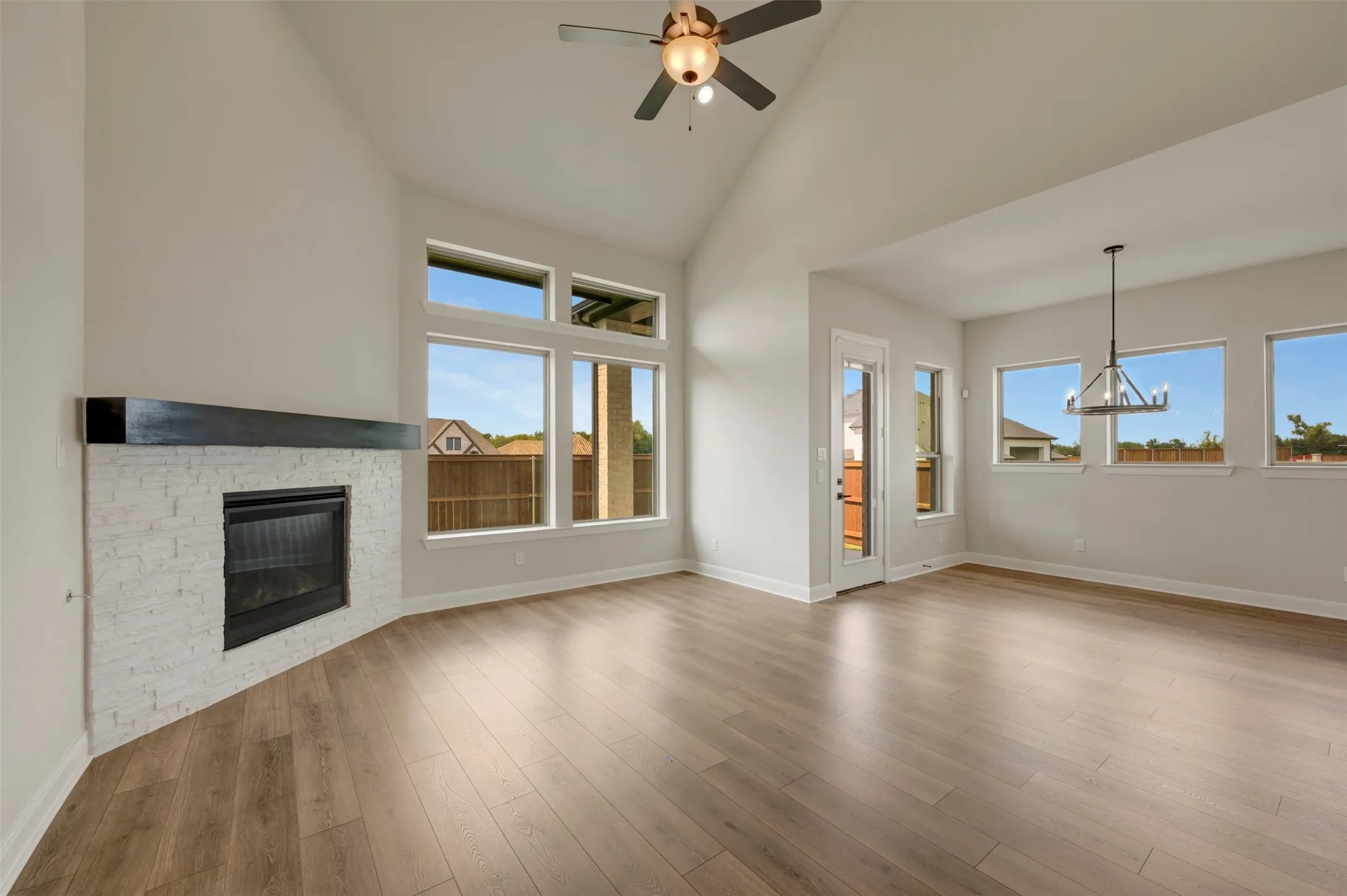 Vaulted ceiling with a ceiling fan, corner fireplace with white stone surround, wood-finish flooring, multiple large windows, and a black chandelier