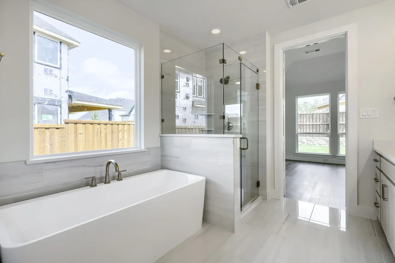 White rectangular soaking tub with brushed nickel fixtures, glass-enclosed shower, white tile flooring, and a large window with white trim