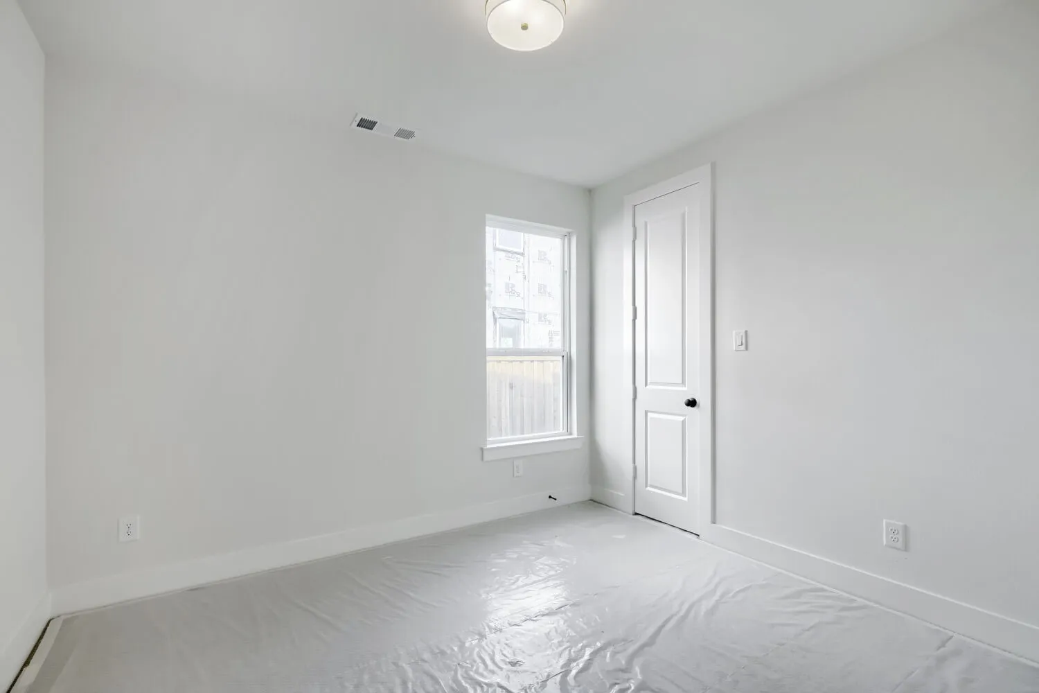 Neutral-toned room featuring a single window with white trim, a flush-mount ceiling light fixture, and a paneled interior door with a black knob