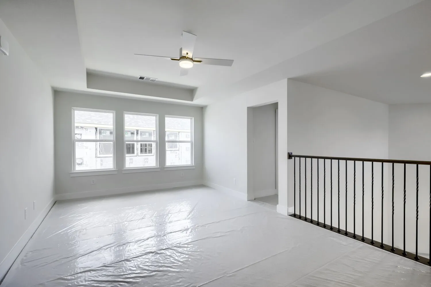 Spacious open area featuring a tray ceiling with a ceiling fan, three large windows, and a black wrought iron railing