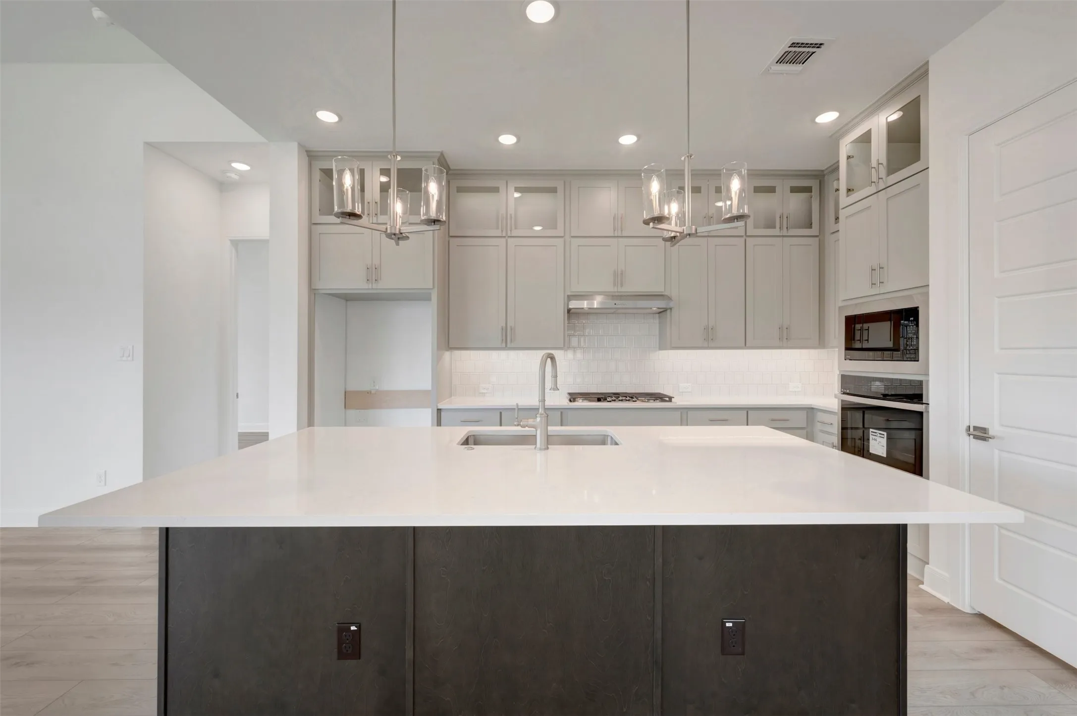 Expansive kitchen island with a light-toned countertop, stainless steel appliances, light gray cabinetry with glass-front uppers, and two pendant light fixtures