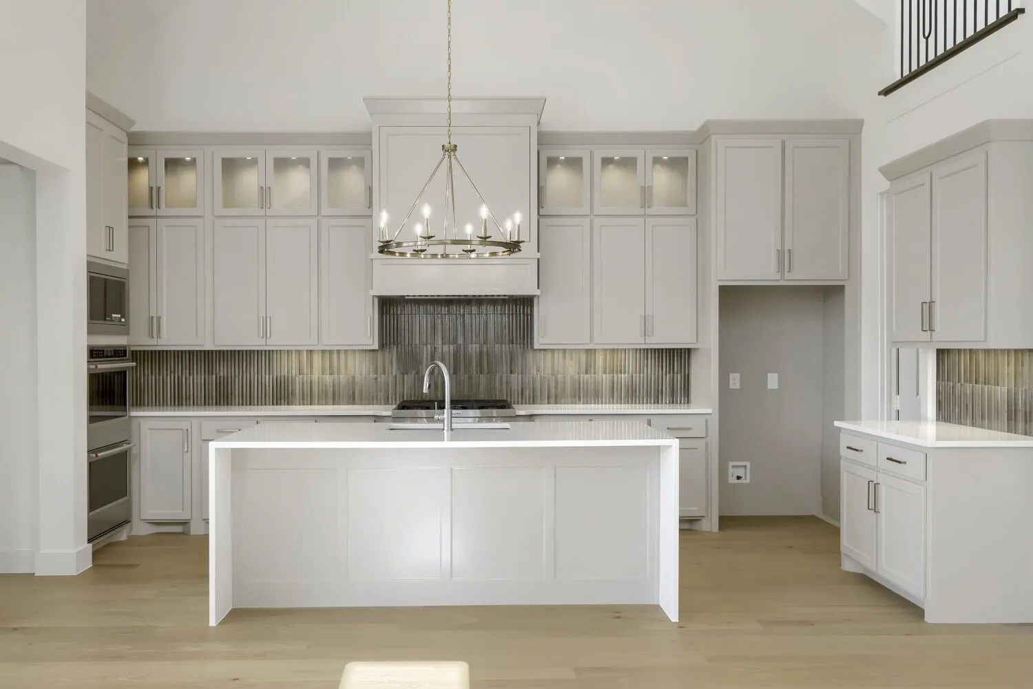 Contemporary kitchen featuring light wood-finish flooring, a large central island with a sink, white cabinetry with under-cabinet lighting, a vertically stacked dark tile backsplash, and a built-in double oven