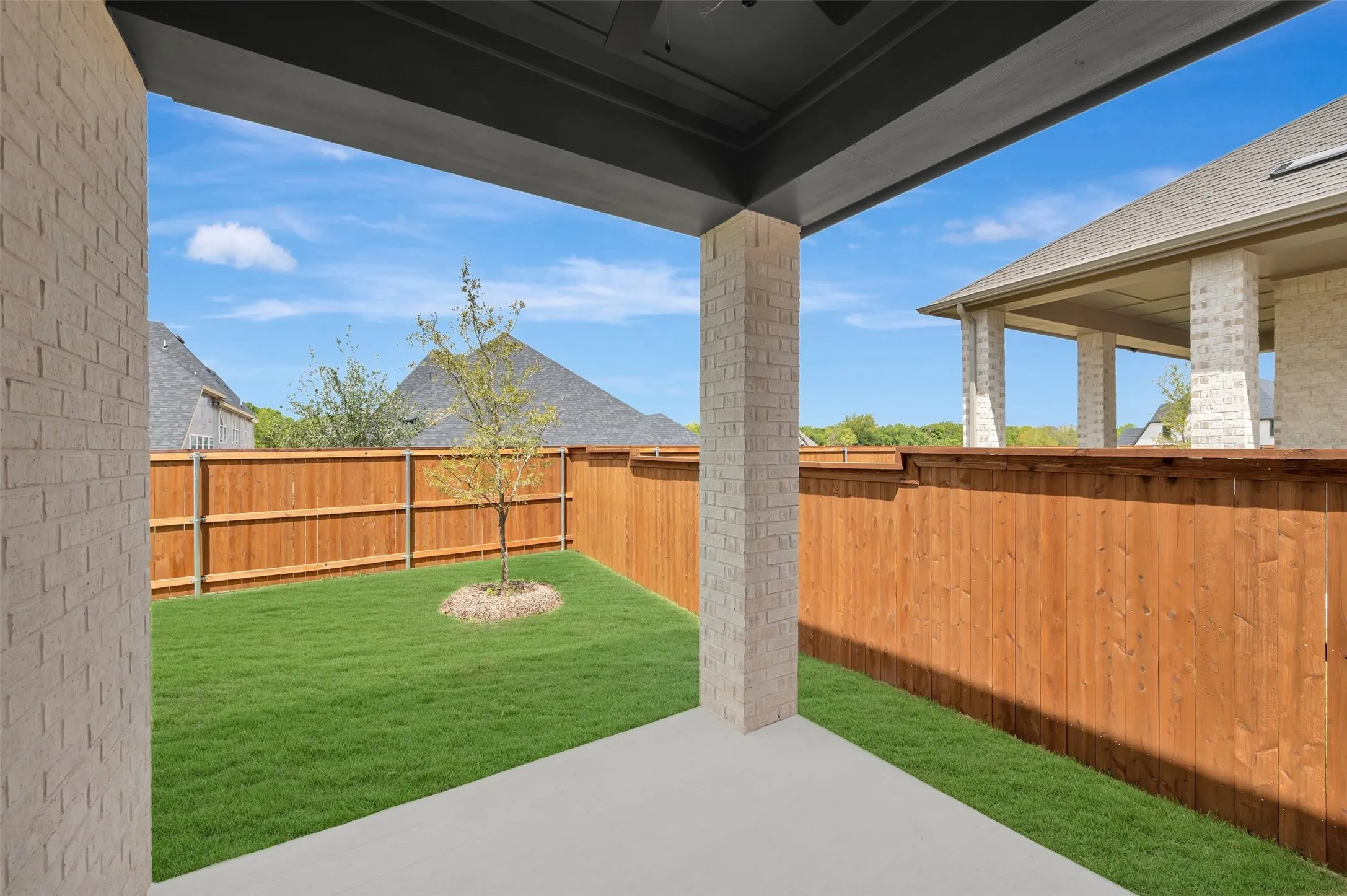 Covered patio with a concrete slab, brick support columns, and a wood fence enclosing a lawn area with a young tree
