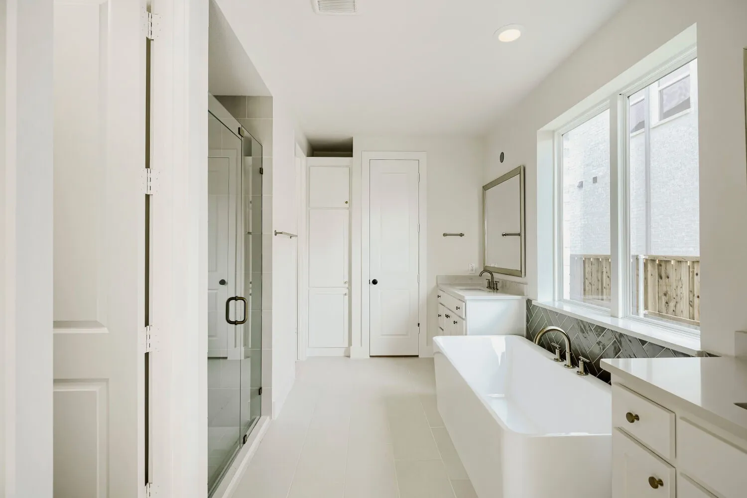 White bathroom featuring a freestanding tub, glass-enclosed shower, dual vanities with undermount sinks, and a herringbone tile accent wall