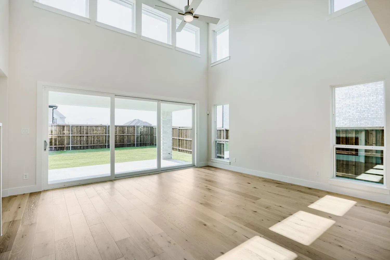 Vaulted ceiling great room with wood-finish flooring, featuring multiple high windows, a ceiling fan, and sliding glass doors opening to a private backyard patio and lawn
