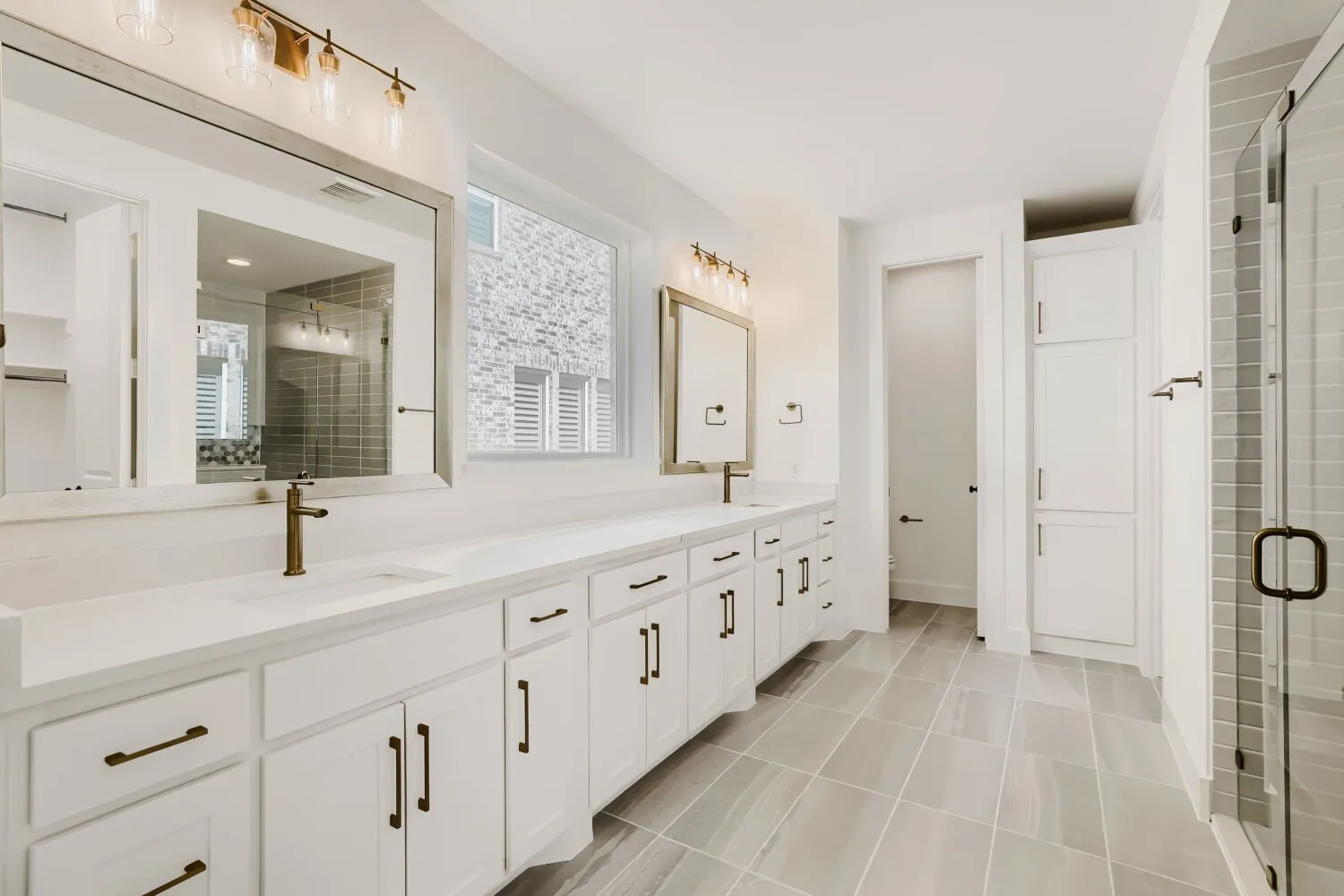 Dual vanity with white cabinetry and integrated sinks, brushed bronze fixtures, and a glass-enclosed shower with tiled walls