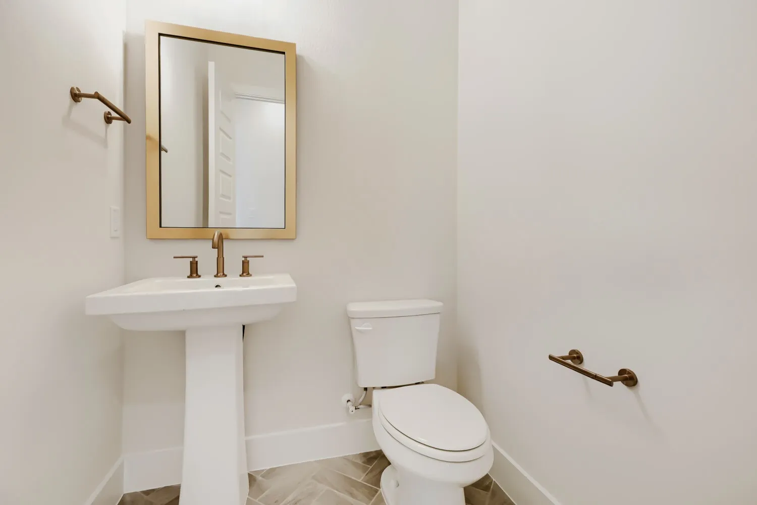 Pedestal sink with brass-finish faucet, rectangular framed mirror, and herringbone pattern tile flooring