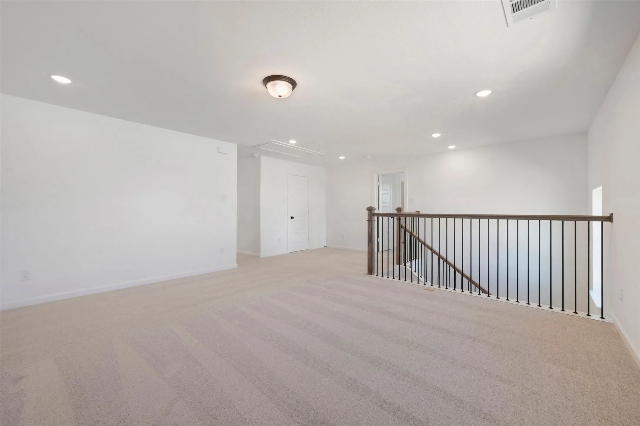 Spacious carpeted landing featuring a dark wood handrail with black metal balusters, recessed lighting, and a flush-mount ceiling fixture