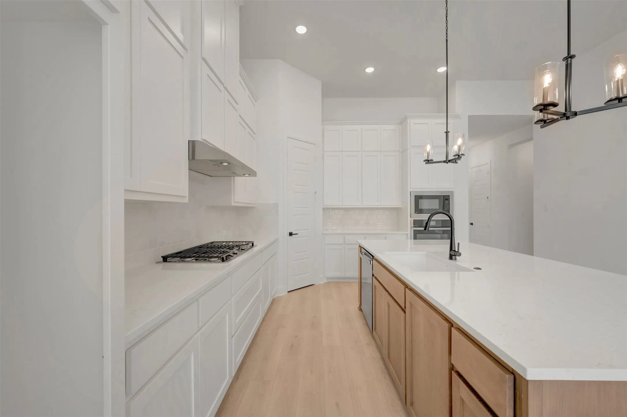 Modern kitchen featuring a large central island with an integrated sink and matte black faucet, stainless steel dishwasher, white countertops, a gas cooktop with range hood, and light wood-finish flooring