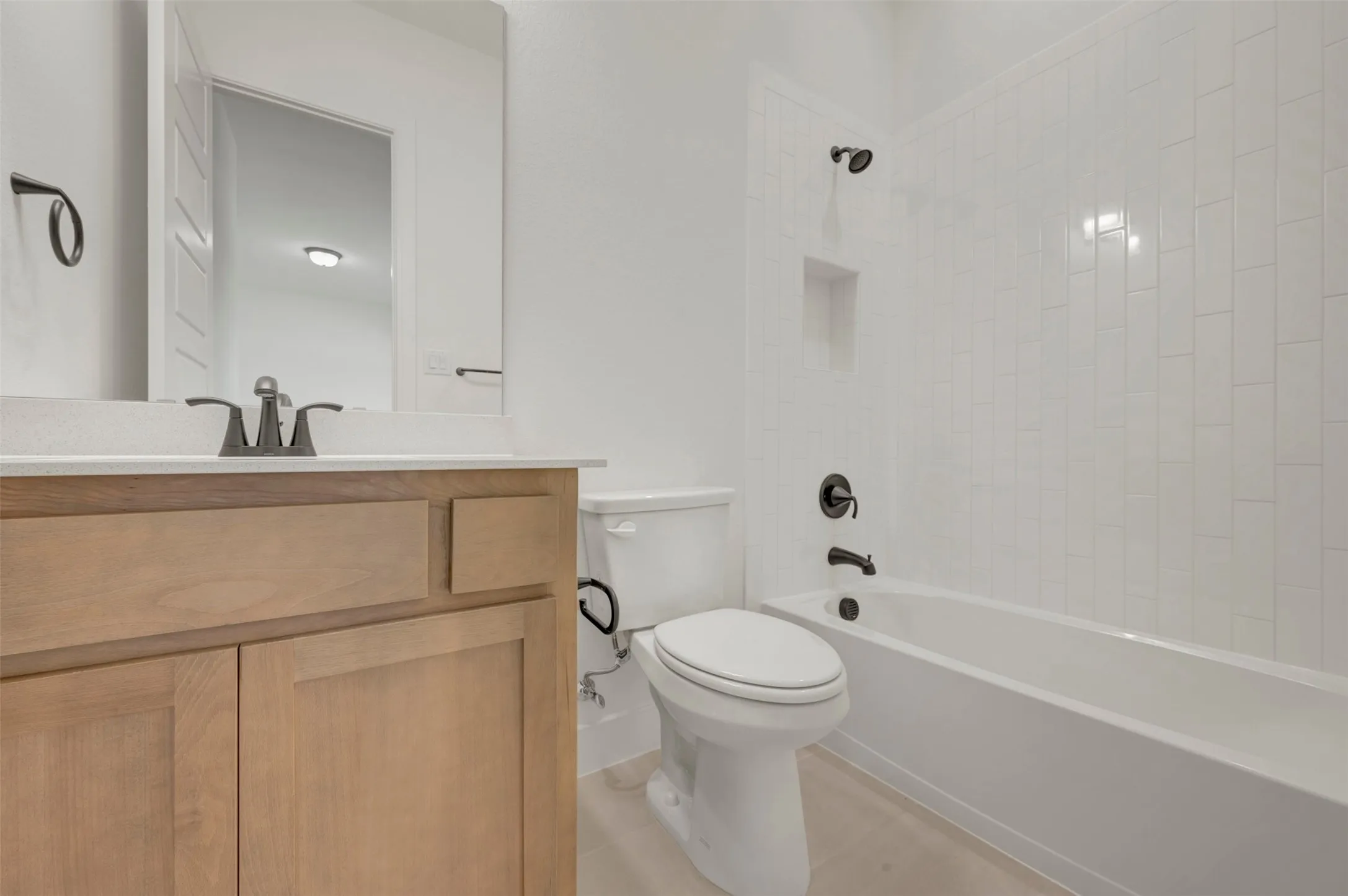 Bathroom featuring a light wood vanity with a white countertop, a rectangular mirror, a white toilet, and a bathtub with white subway tile surround and a recessed niche