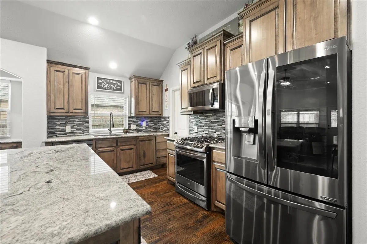 Kitchen featuring stainless steel appliances, vaulted ceiling, light stone counters, wood finish cabinetry, and tasteful backsplash