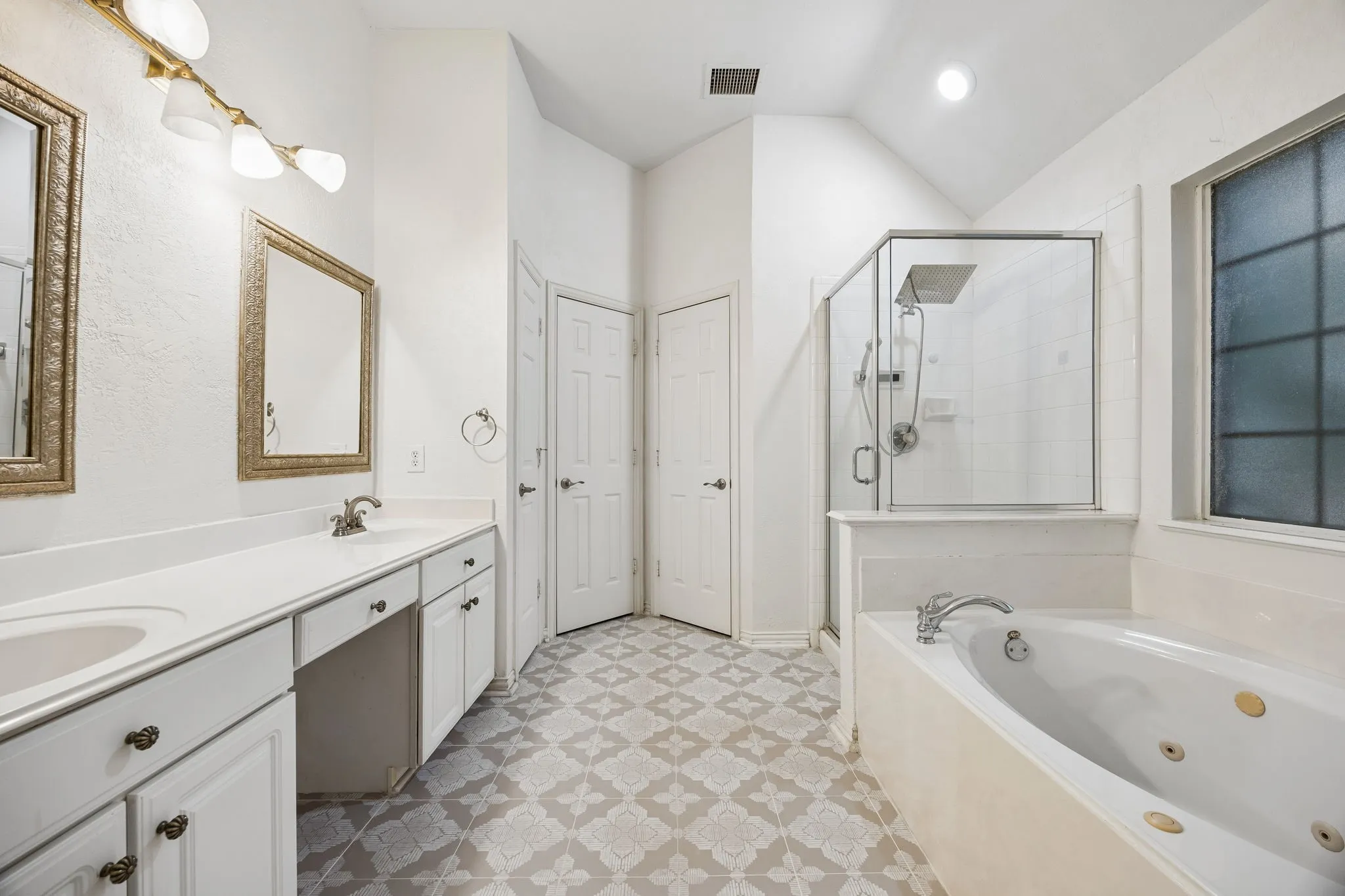 Bathroom featuring a dual vanity with white cabinetry and integrated sinks, a built-in jetted tub, a glass-enclosed shower with a rainfall showerhead, patterned floor tiling, and a vaulted ceiling