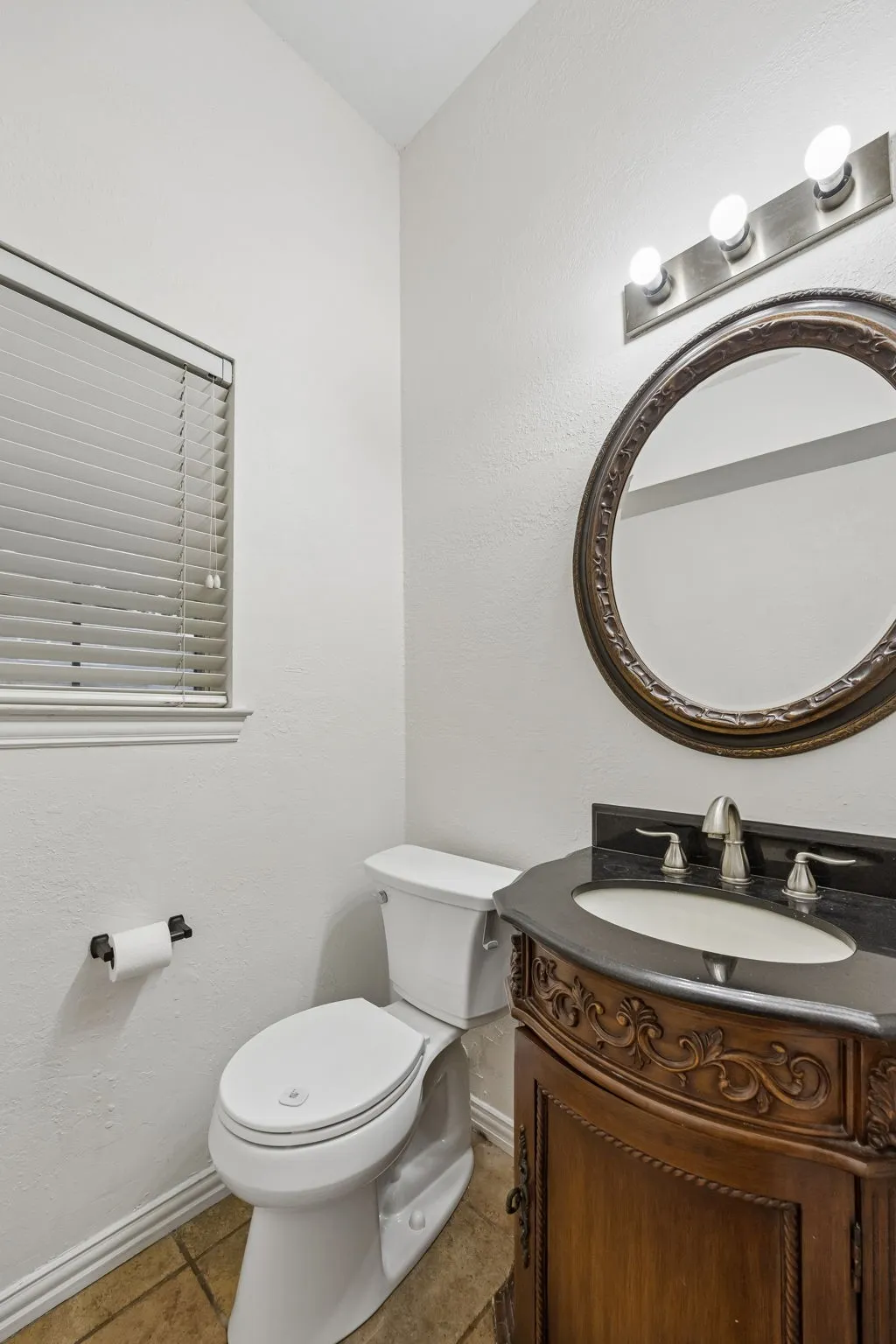 Powder room featuring a single-pane window with blinds, a white toilet, an ornate wood-finish vanity with a dark countertop and undermount sink, a chrome faucet, and a decorative oval mirror under a three-bulb light fixture