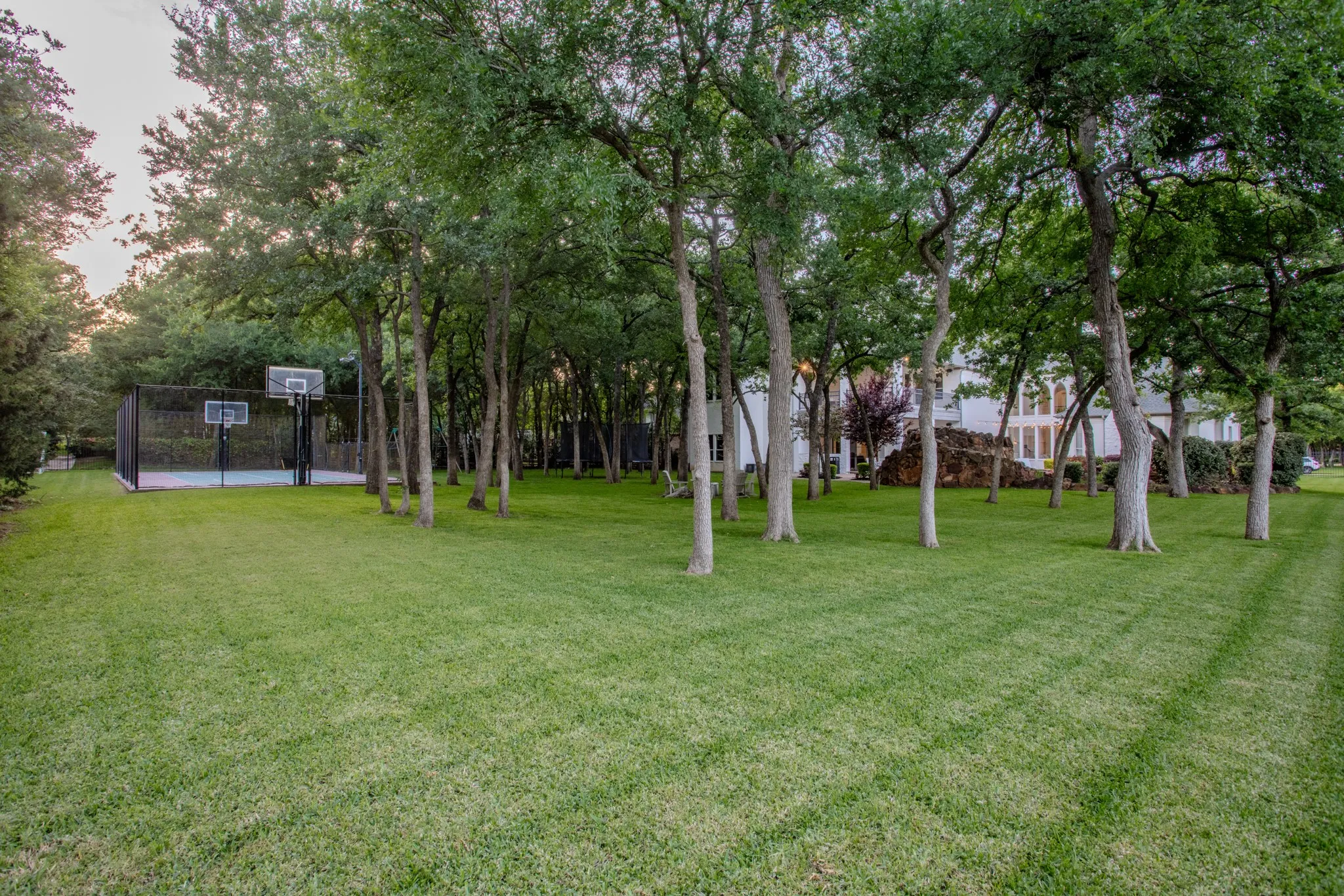 Expansive grassy yard featuring an enclosed sport court with two basketball hoops, mature trees, and a distant property exterior with arched windows