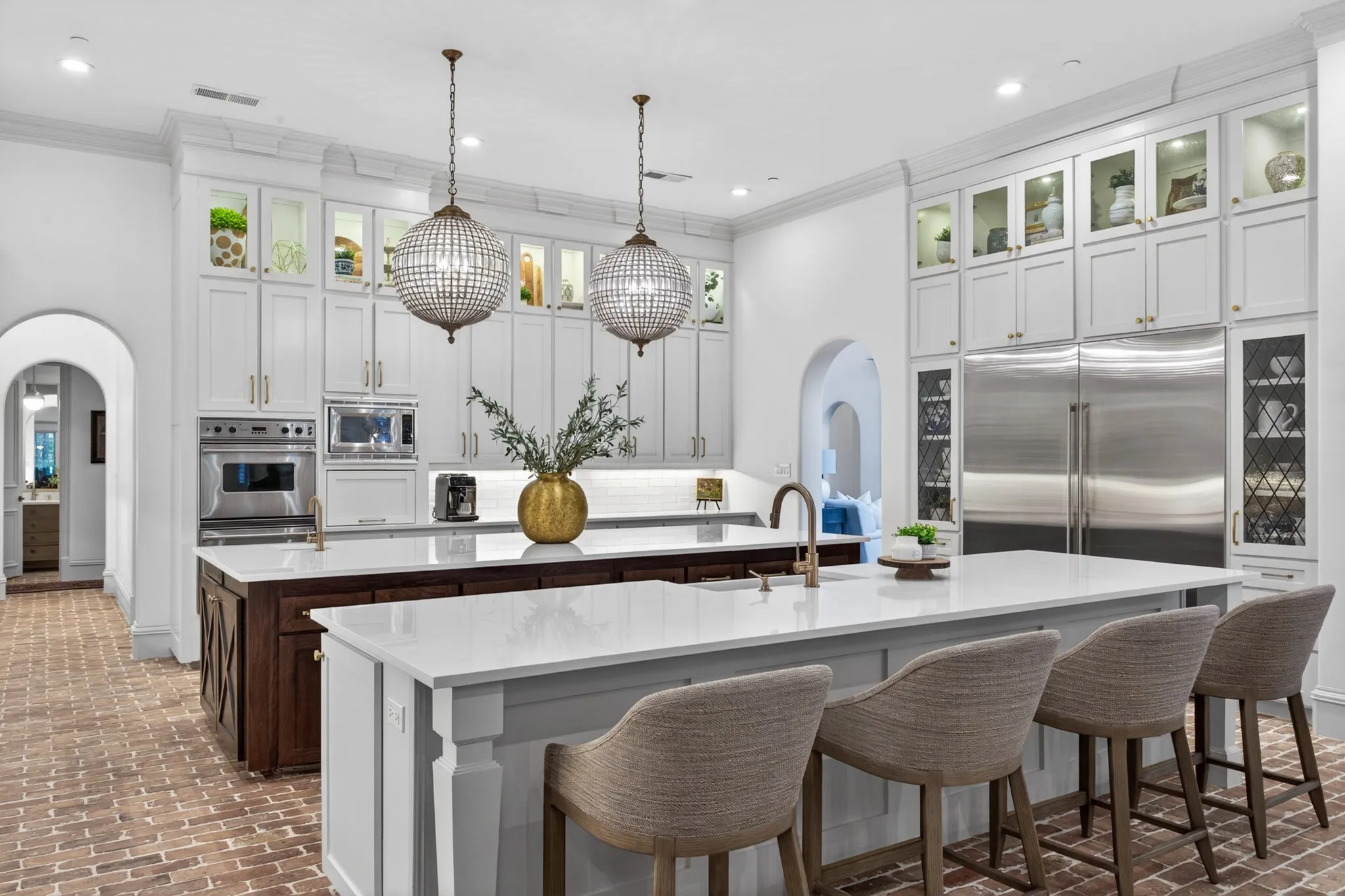 Gourmet kitchen featuring a brick-finish flooring, dual islands with white countertops, integrated stainless steel appliances, white cabinetry with glass-front uppers, and two ornate pendant lights