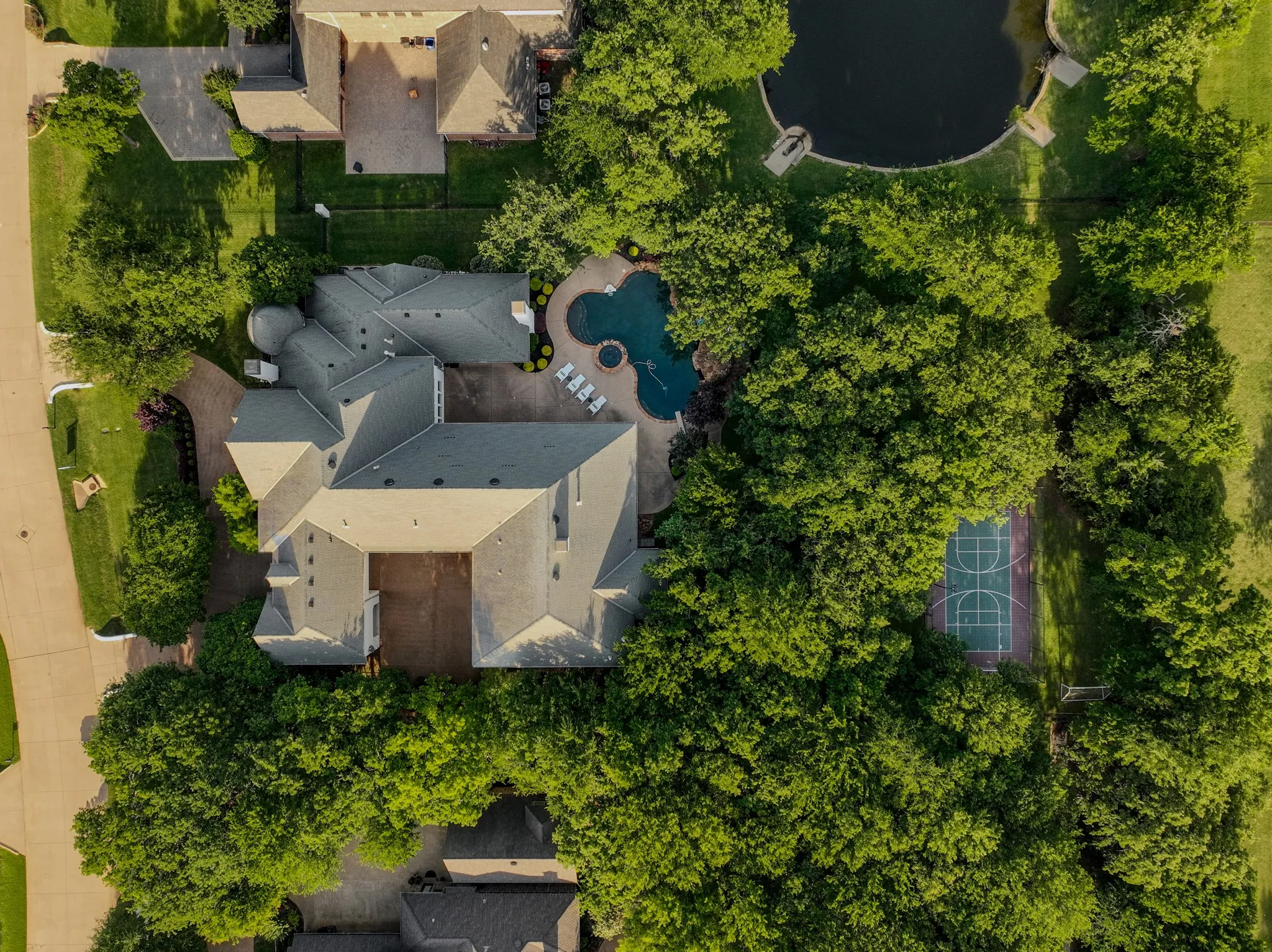 Aerial estate overview showcasing a multi-wing residence, a freeform swimming pool with integrated spa, a dedicated sports court, mature tree landscaping, and a nearby pond