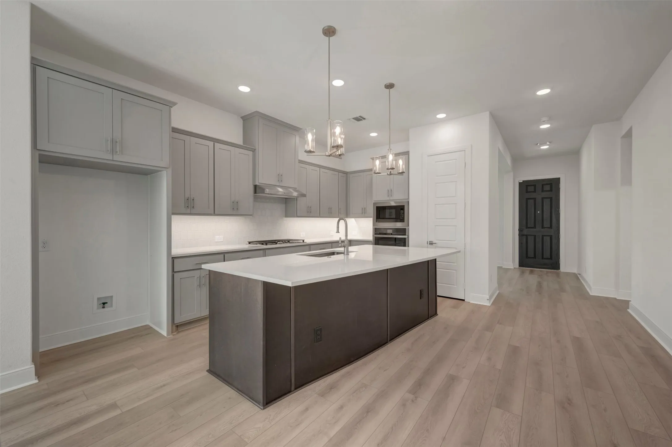 Open-concept kitchen featuring an oversized island with an undermount sink, light gray cabinetry, stainless steel appliances, and wood-finish flooring