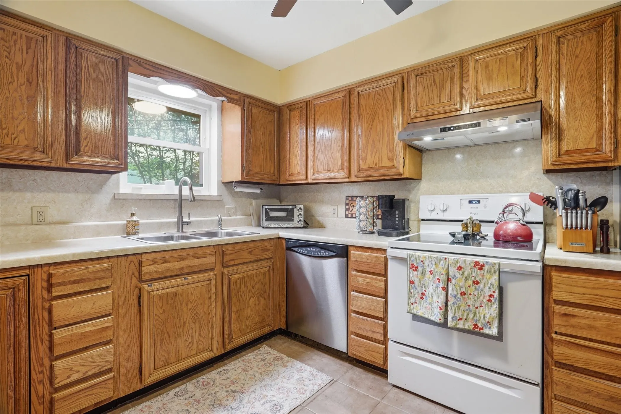 Kitchen featuring white electric range oven, wood finish cabinets, light countertops, stainless steel dishwasher, and light tile patterned flooring