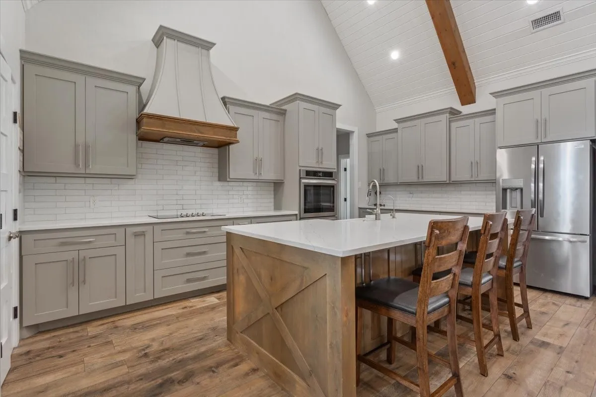Kitchen with gray cabinetry, stainless steel appliances, dark wood-type flooring, light stone counters, and a center island with sink