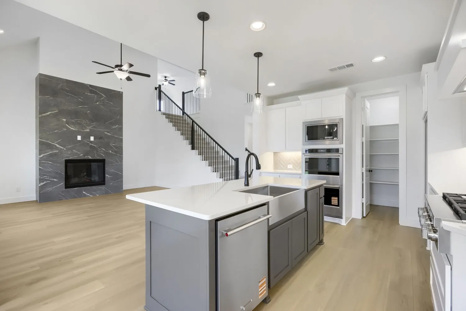 Open-concept living space featuring a central kitchen island with a farmhouse sink, stainless steel appliances, a pantry with built-in shelving, a fireplace with a stone surround, and wood-finish flooring throughout