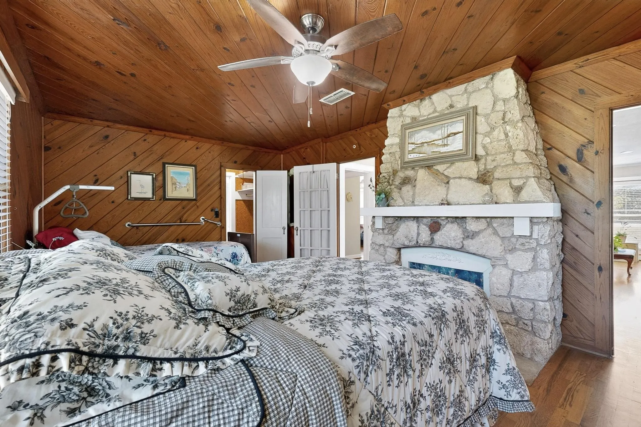 Bedroom featuring wooden walls, wood finished floors, a ceiling fan, wooden ceiling, and a stone fireplace