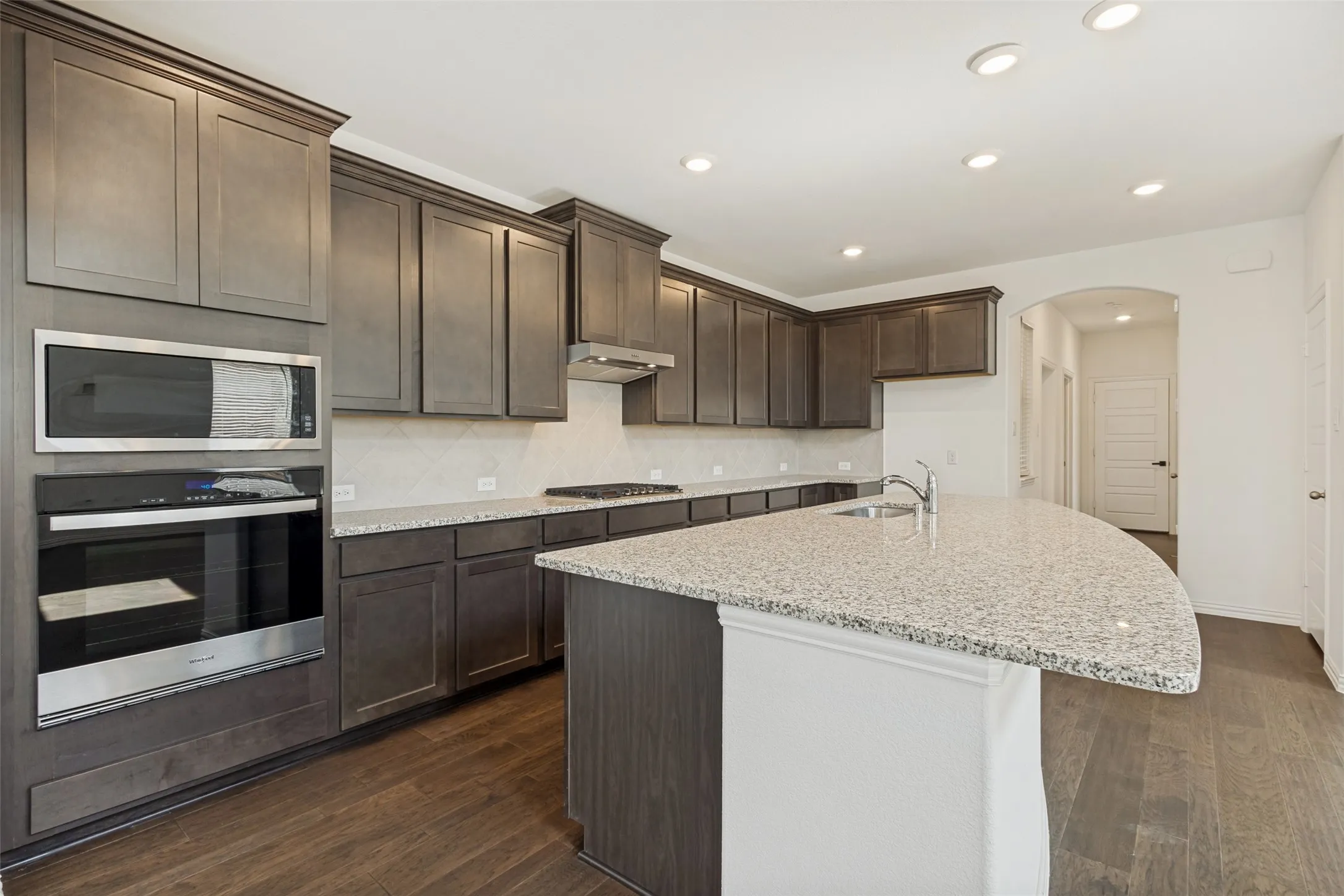 Kitchen featuring dark wood-finish cabinetry, light speckled countertops, a built-in stainless steel microwave and oven, a gas cooktop, and wood-finish flooring