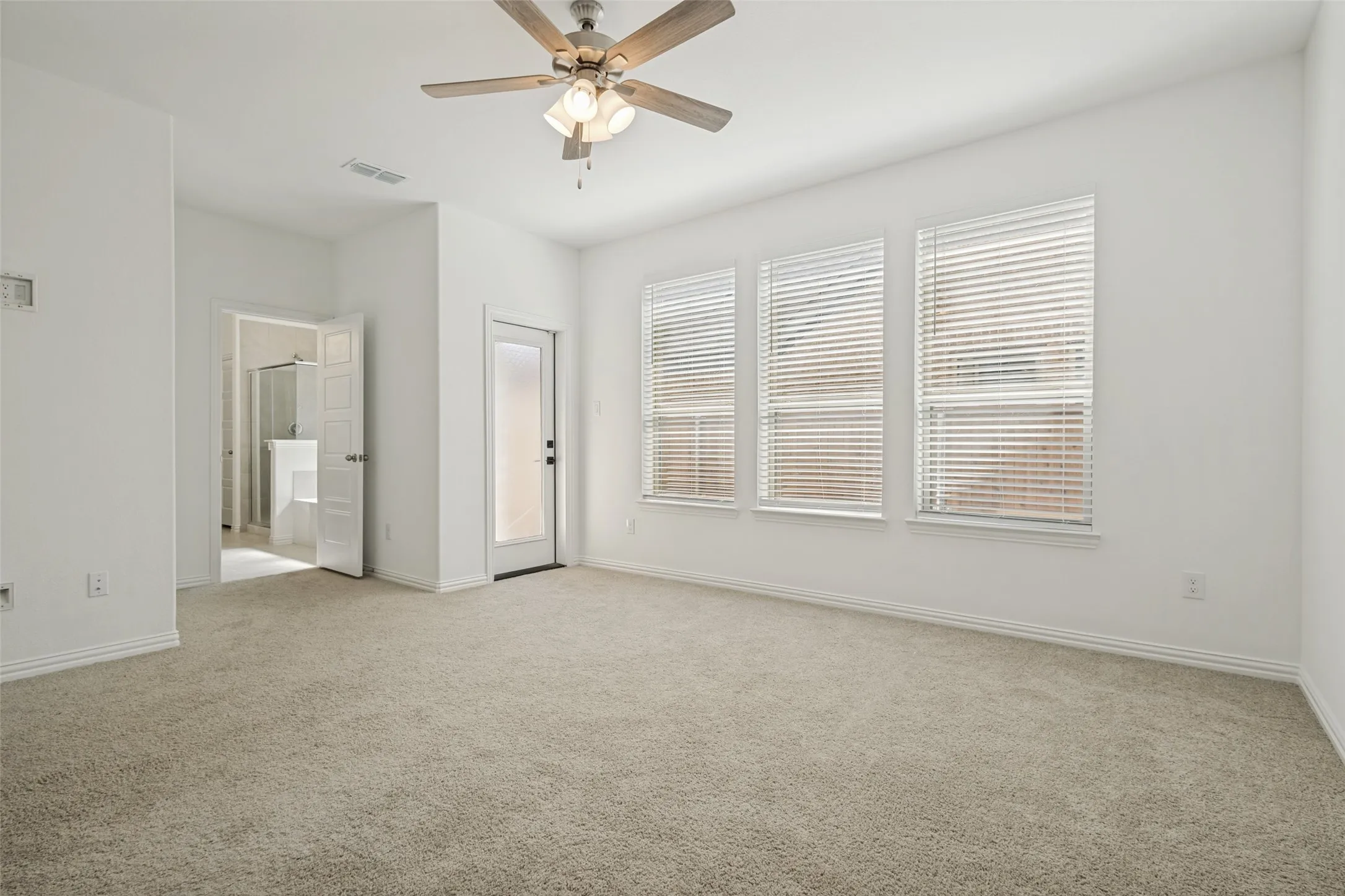 Spacious carpeted room featuring a ceiling fan with light kit, three large windows with blinds, a solid door leading to an outdoor space, and an open doorway to an en-suite bathroom with a glass-enclosed shower